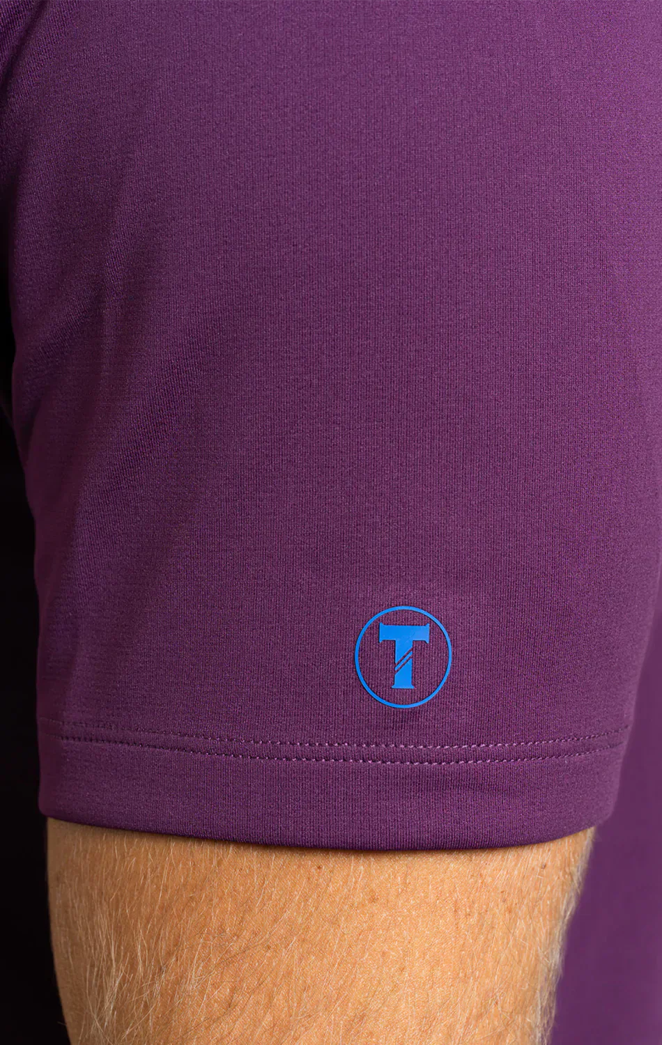 Close-up of a person’s arm in the Twillory Short Sleeve Polo Solid // Plum, highlighting the moisture-wicking sleeve with a small blue circular logo featuring a capital T. Background and other details are not visible.