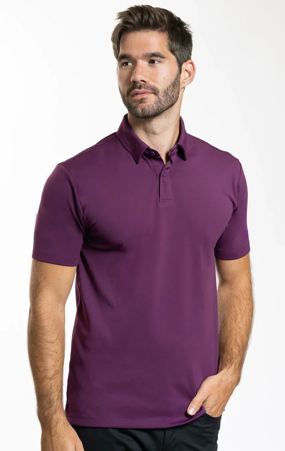 A man with short brown hair and a beard wears the Twillory Short Sleeve Polo Solid in Plum, standing against a white background, looking slightly left with a neutral expression and one hand in his pocket.
