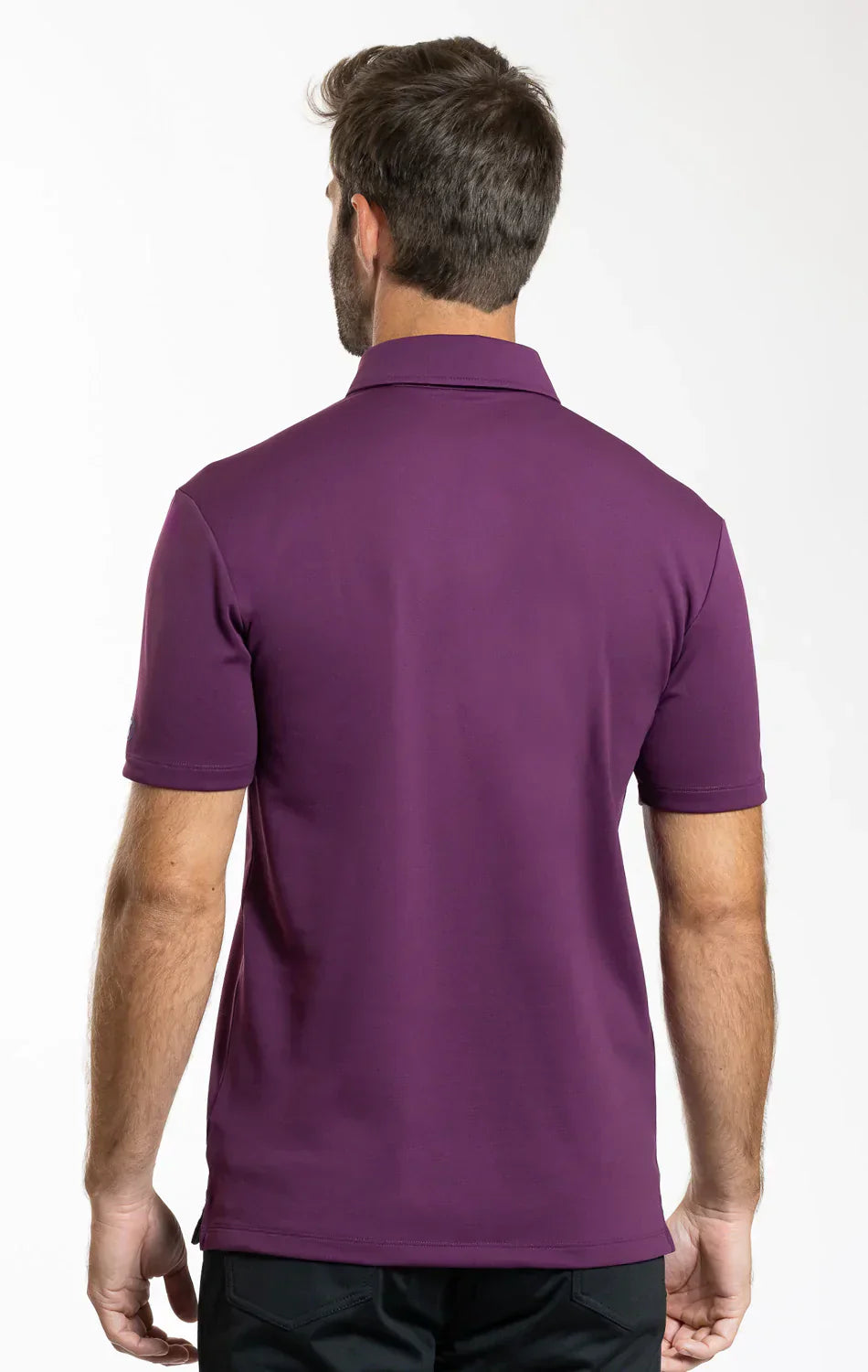 A man with short brown hair stands with his back to the camera, wearing Twillory's Short Sleeve Polo Solid in Plum, featuring a dress collar and black pants, set against a plain white background.