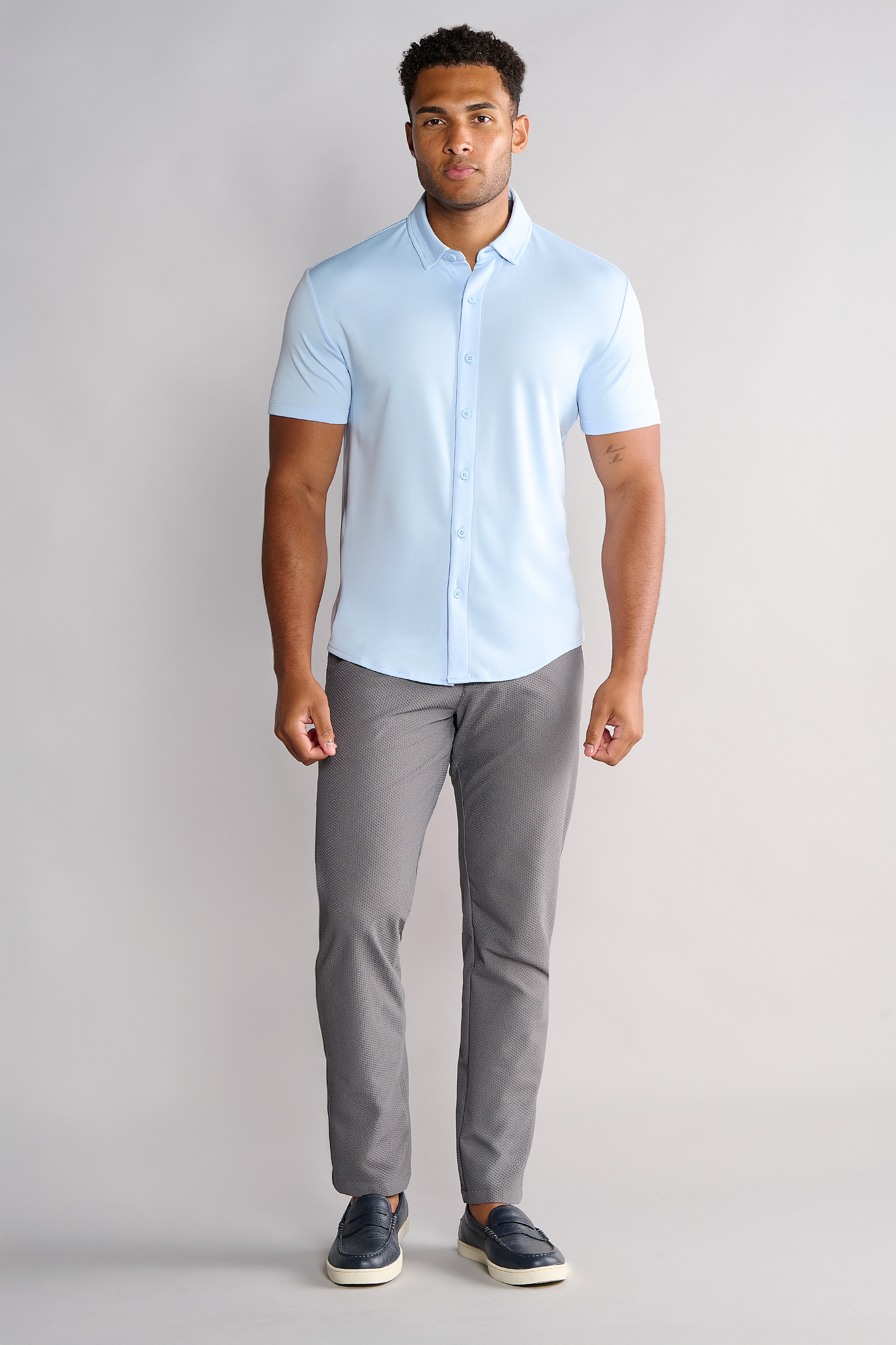 A man stands facing forward against a plain light background, wearing a Twillory Short Sleeve Button Down Polo Solid in Powder Blue, gray slim-fit pants, and dark slip-on shoes. His arms are relaxed at his sides and he has a neutral expression.