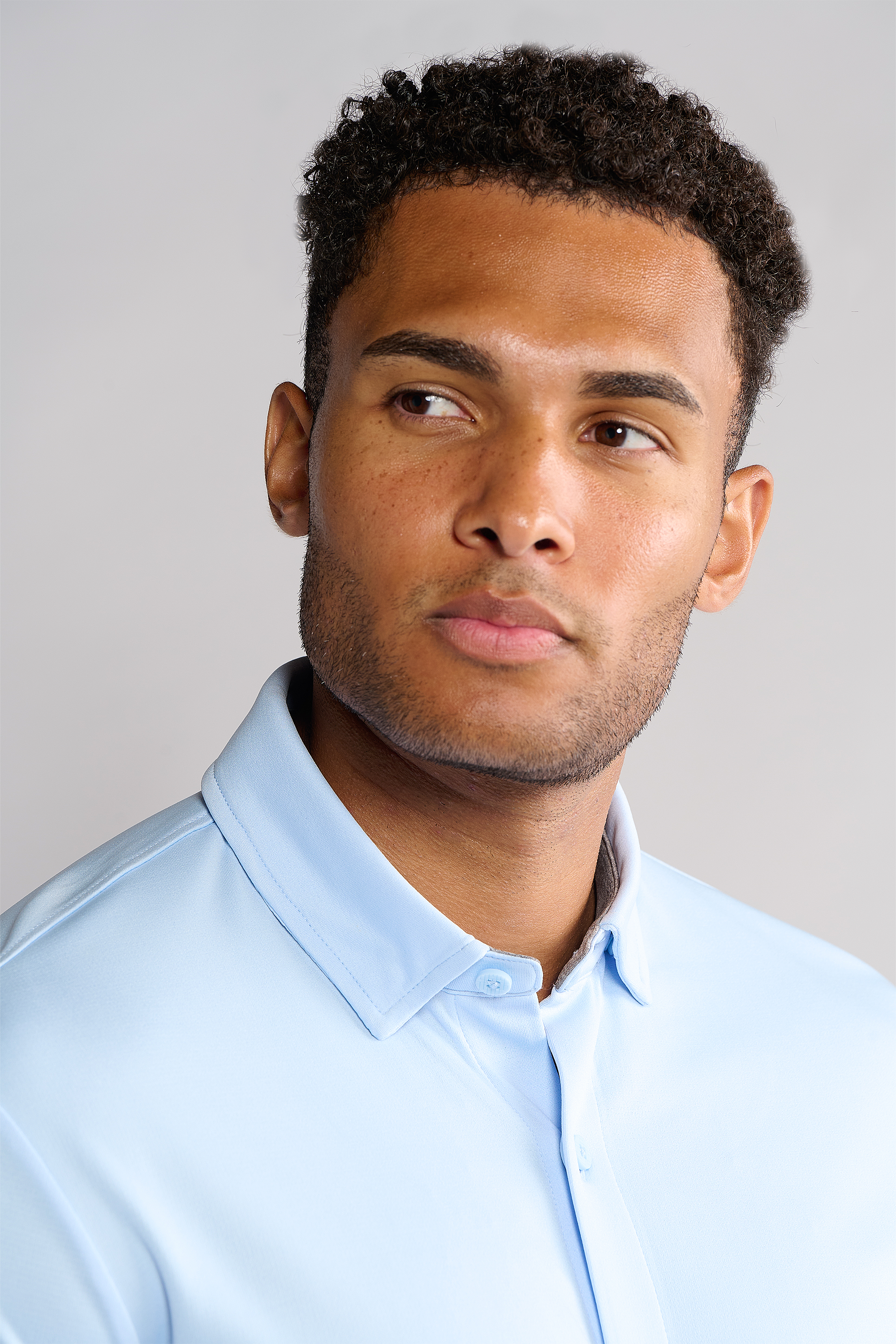 A young man with short curly hair and a trimmed beard looks to the side, wearing Twillory’s Short Sleeve Button Down Polo Solid in Powder Blue. The plain background highlights his thoughtful expression.
