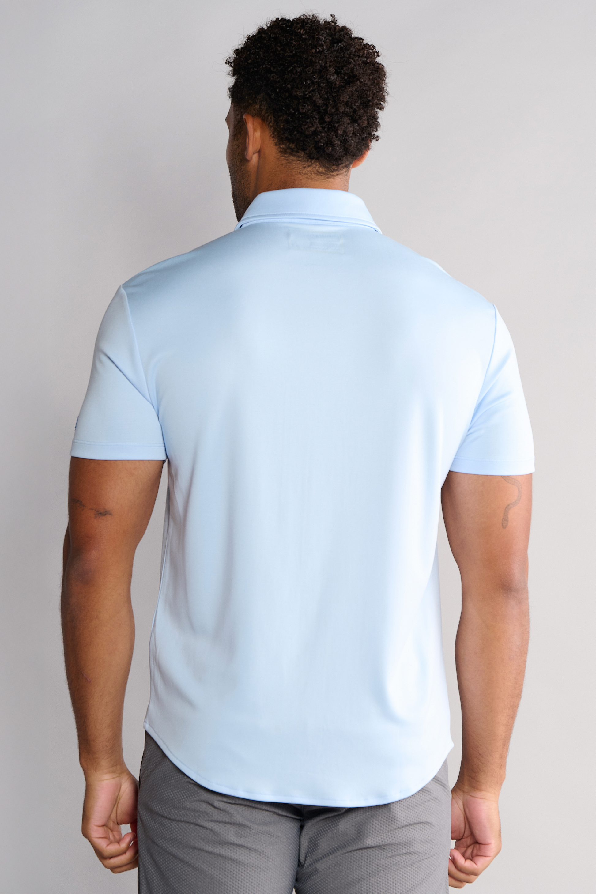 A man with short curly hair faces away, wearing a Twillory Short Sleeve Button Down Polo Solid in Powder Blue and dark gray pants. His arms are relaxed at his sides, revealing a small tattoo on his left upper arm. The background is plain light gray.
