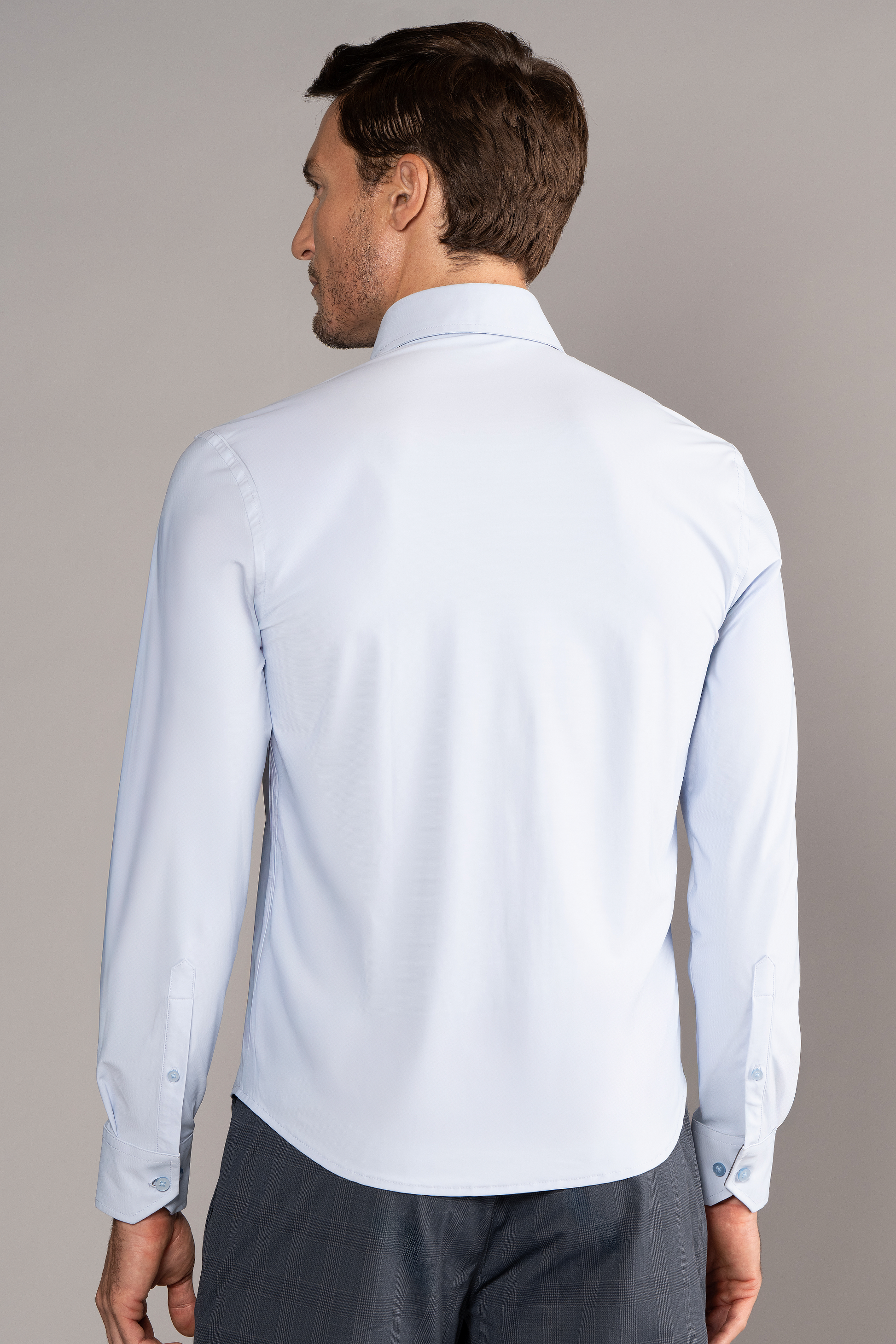 A man faces away from the camera, wearing Twillory’s The Event Shirt // Powder Blue—an office shirt in wrinkle-free stretch, warp-knit tech fabric with a classic collar and buttoned cuffs. A plain gray backdrop highlights the attire.