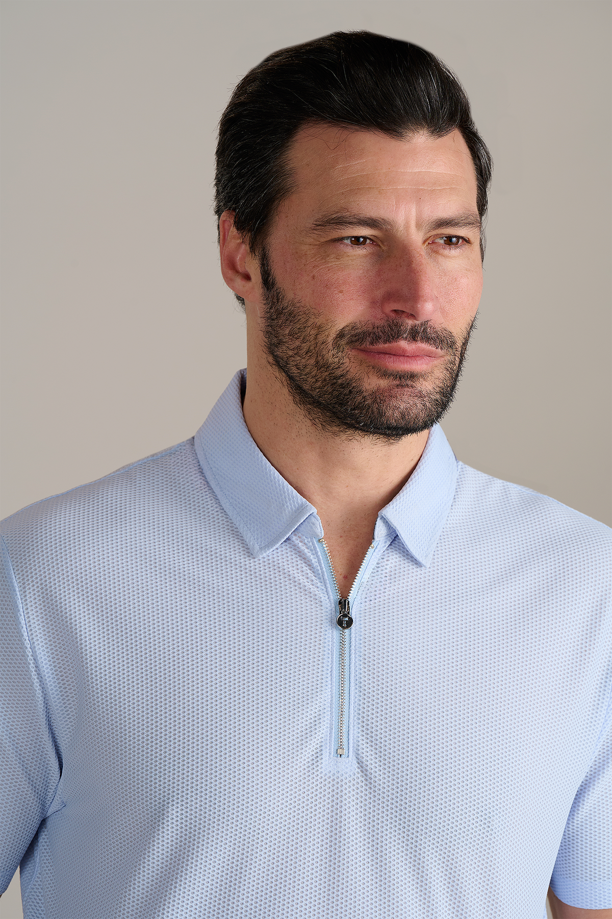 A man with dark hair and a trimmed beard wears the Twillory AIR Zip Polo Solid in Powder Blue, featuring short sleeves and a zip collar. He faces right, smiling softly against a plain background that keeps the focus on him.