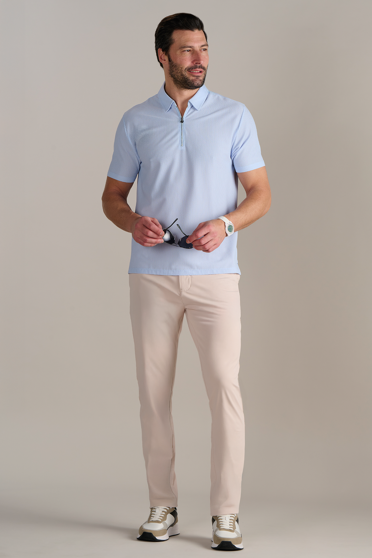 A man with short brown hair and a trimmed beard stands facing forward, holding sunglasses. He wears the Twillory AIR Zip Polo Solid in Powder Blue, beige pants, a white watch, and white sneakers against a plain background.