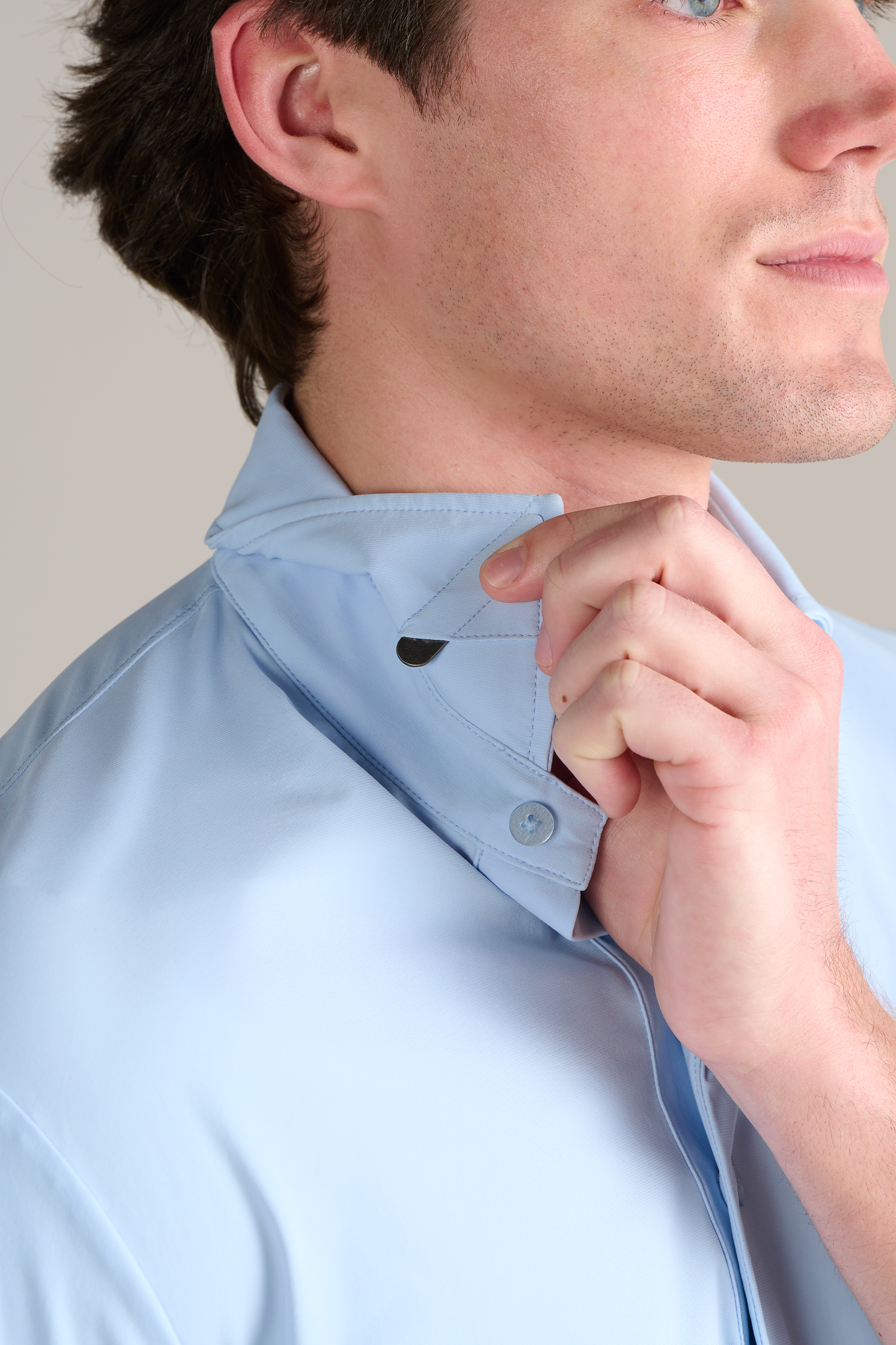 A person lifts the collar of a Twillory Event Polo Solid in Powder Blue with their right hand, revealing a hidden black collar stay for added structure. Their face is partly visible, showing light skin and short brown hair.