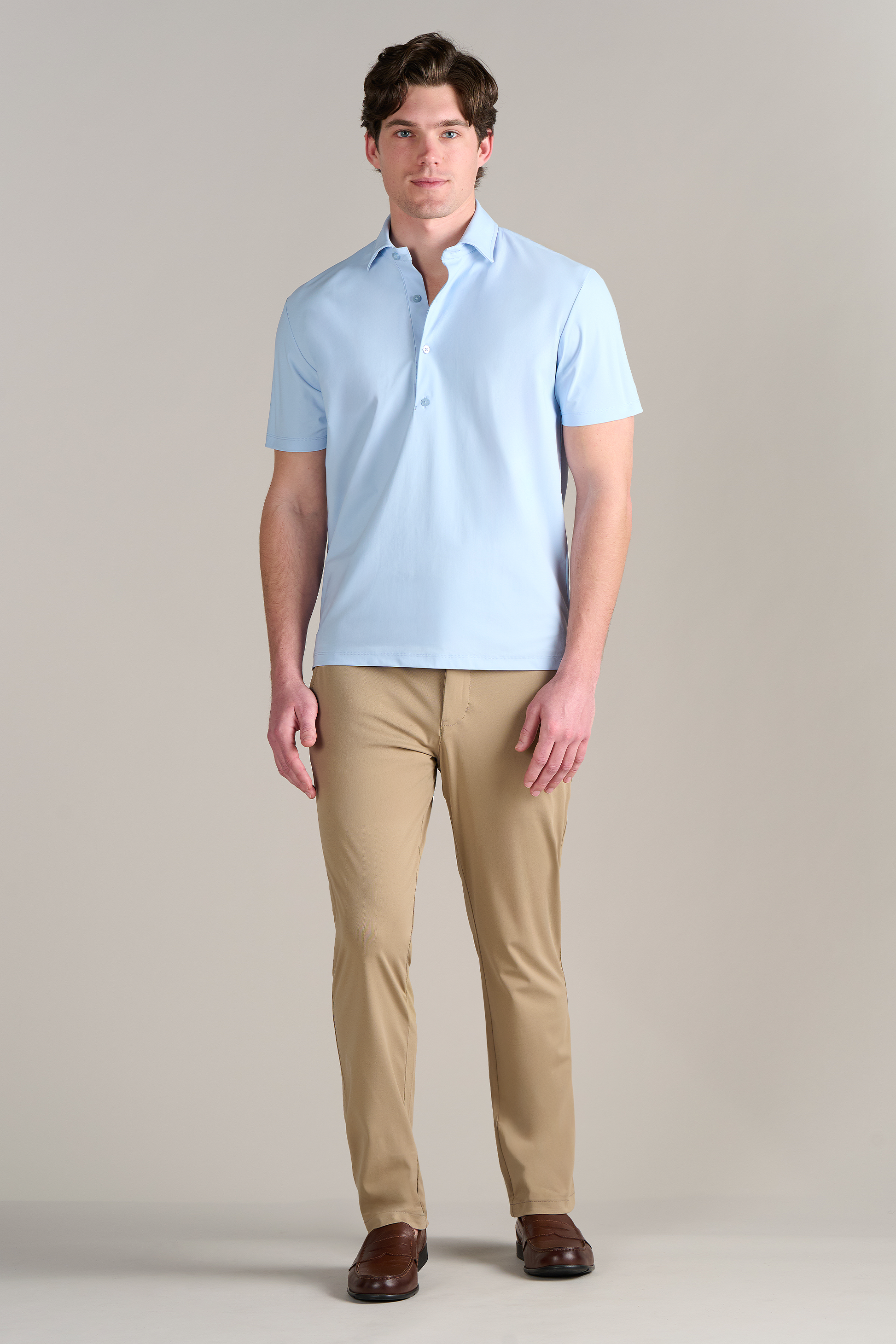 A man with short brown hair and a neutral expression stands against a plain background wearing Twillory’s Event Polo Solid in Powder Blue, beige slim-fit pants, and brown loafers. His arms are relaxed at his sides.