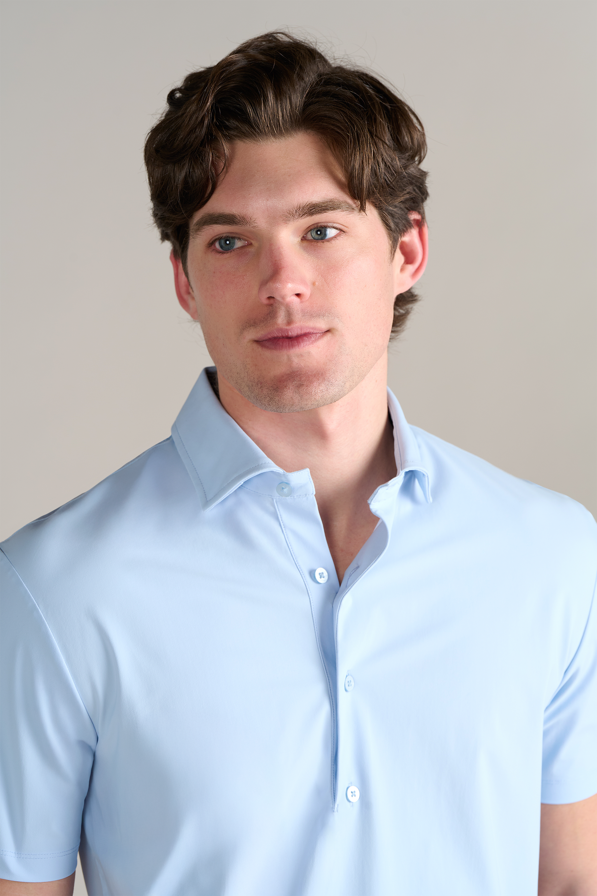 A young man with wavy brown hair and blue eyes wears the Twillory Event Polo Solid in Powder Blue, a light blue short-sleeve shirt with a dress collar made from wrinkle-free, performance stretch fabric, standing against a plain light background.