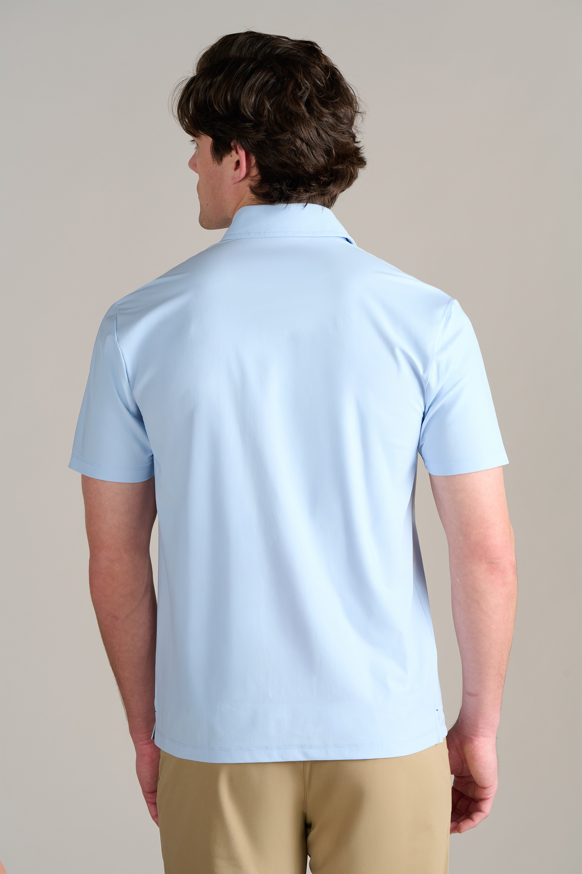 A man is seen from behind wearing Twillory's Event Polo Solid in Powder Blue, featuring a dress shirt collar, and khaki pants. He has short wavy brown hair and stands against a light background with his arms at his sides.