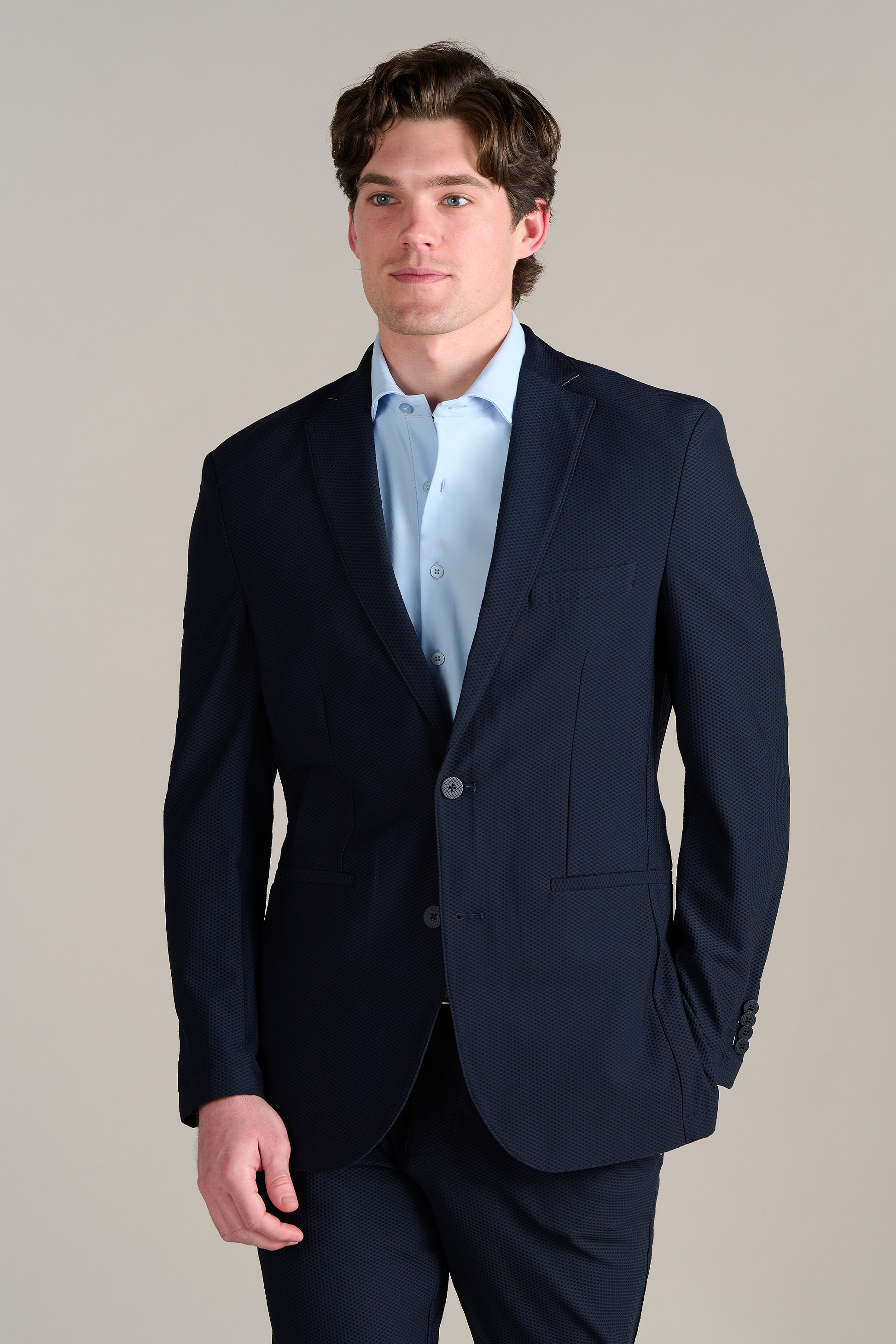 A man with wavy brown hair models the Twillory Event Polo Solid in Powder Blue, standing slightly left with one hand by his side and the other in his pocket against a plain light gray background.