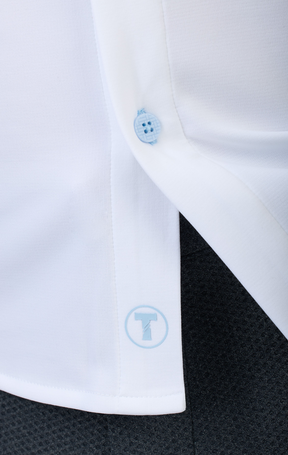 A close-up of Twillory's Long Sleeve Button Down Polo Contrast in Powder Blue White shows its crisp hem, subtle blue logo, light blue button, and fine stitching over dark pants—highlighting moisture-wicking fabric and a discreet branded graphic.