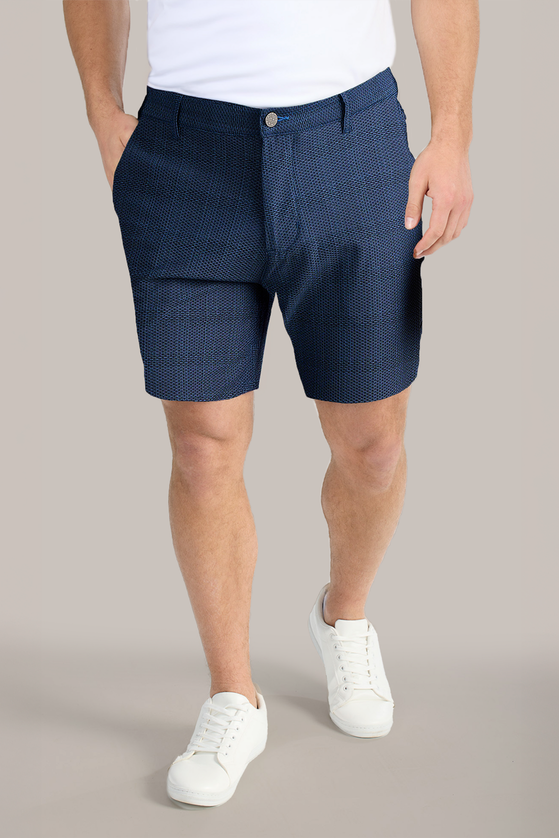 A person stands with hands in the pockets of Twillory AIR Shorts in dark blue pattern, paired with a white T-shirt and white sneakers. The plain background highlights this casual summer outfit. The face is not visible.