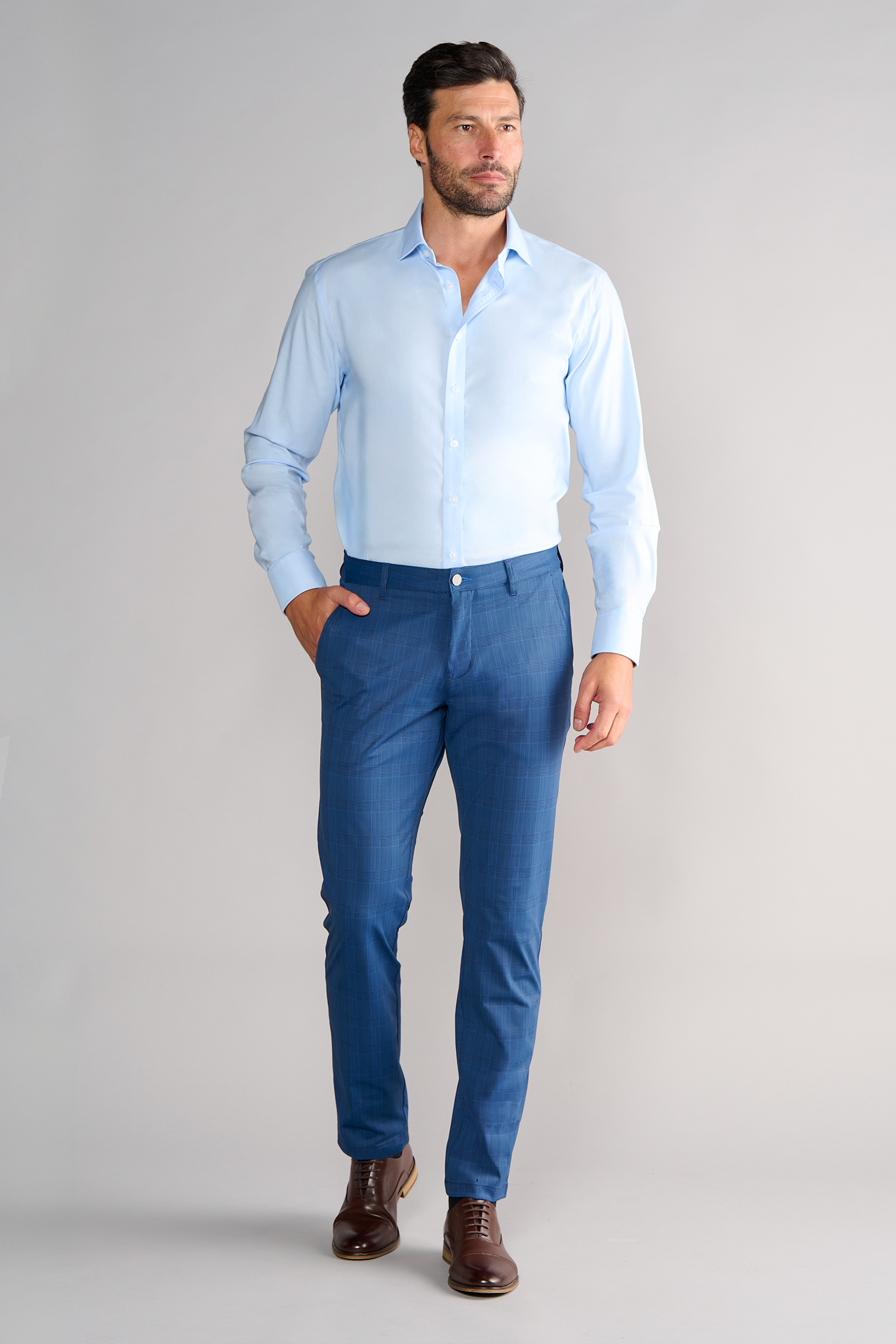 A man with short dark hair and a beard wears Twillory’s Performance Pants in Blue Prince of Wales, paired with a light blue shirt and brown dress shoes. He stands against a plain light gray background, looking slightly to the side with one hand in his pocket.
