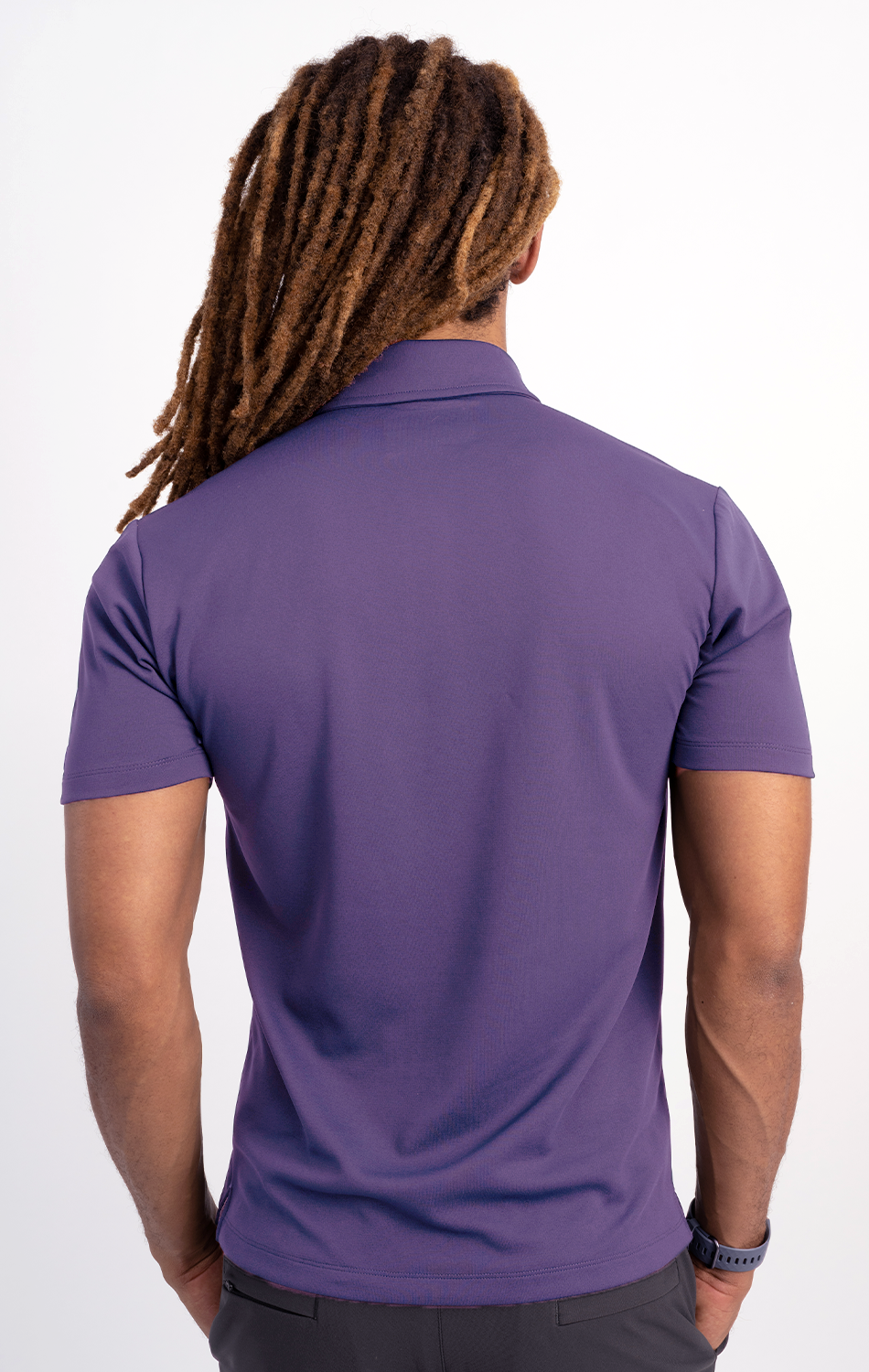 A person with long, brown dreadlocks is shown from the back wearing the Twillory Short Sleeve Polo Solid in Purple and dark pants. Their arms are relaxed at their sides against a plain white background.