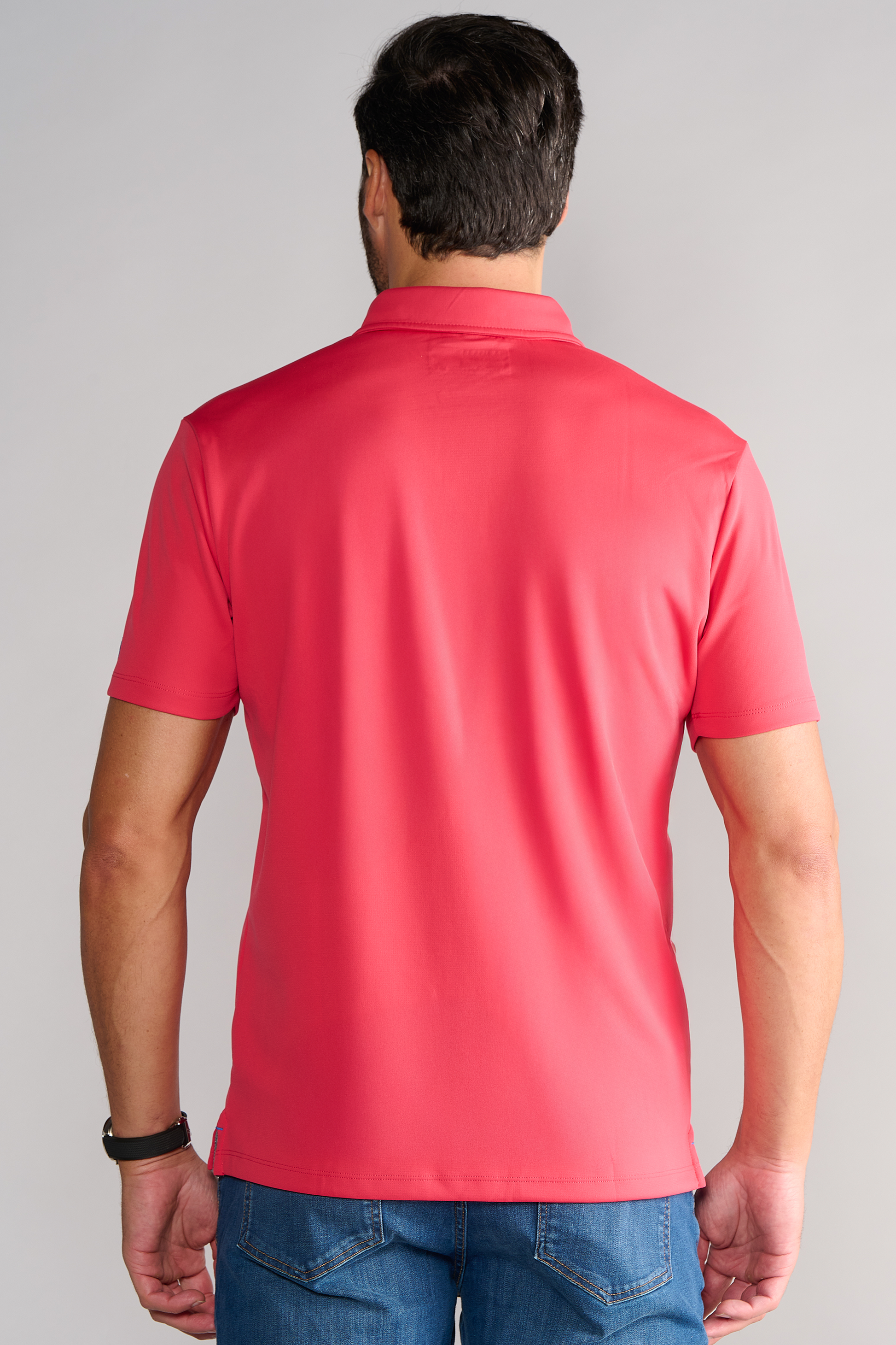 A man with short dark hair is shown from behind wearing the Twillory Short Sleeve Polo Solid in red and blue jeans. He sports a black watch on his left wrist. The wrinkle-free shirt pops against a plain, light gray background.