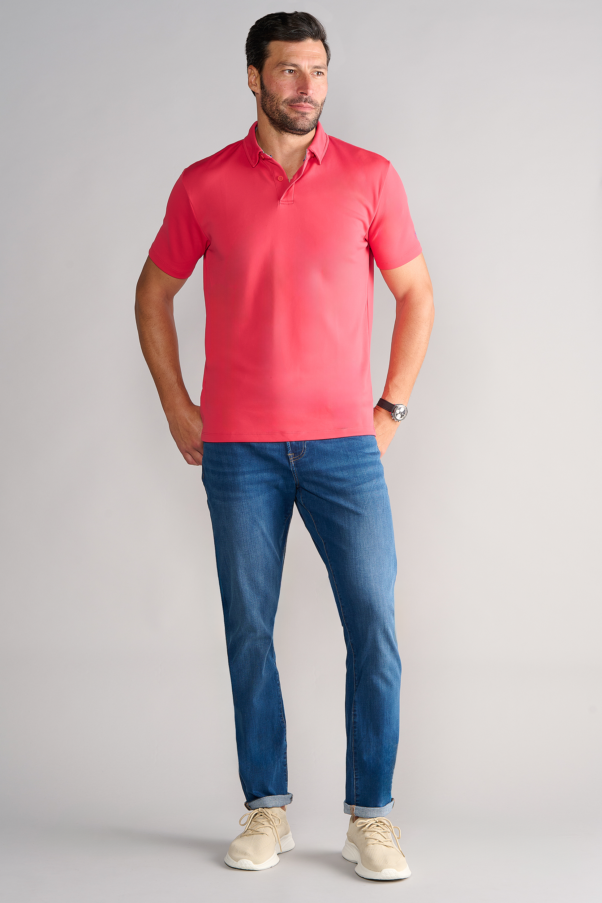 A man with short dark hair and a beard stands against a plain light gray background, wearing the Twillory Short Sleeve Polo Solid in Red, blue jeans, beige sneakers, and a wristwatch. His hands are relaxed at his sides.