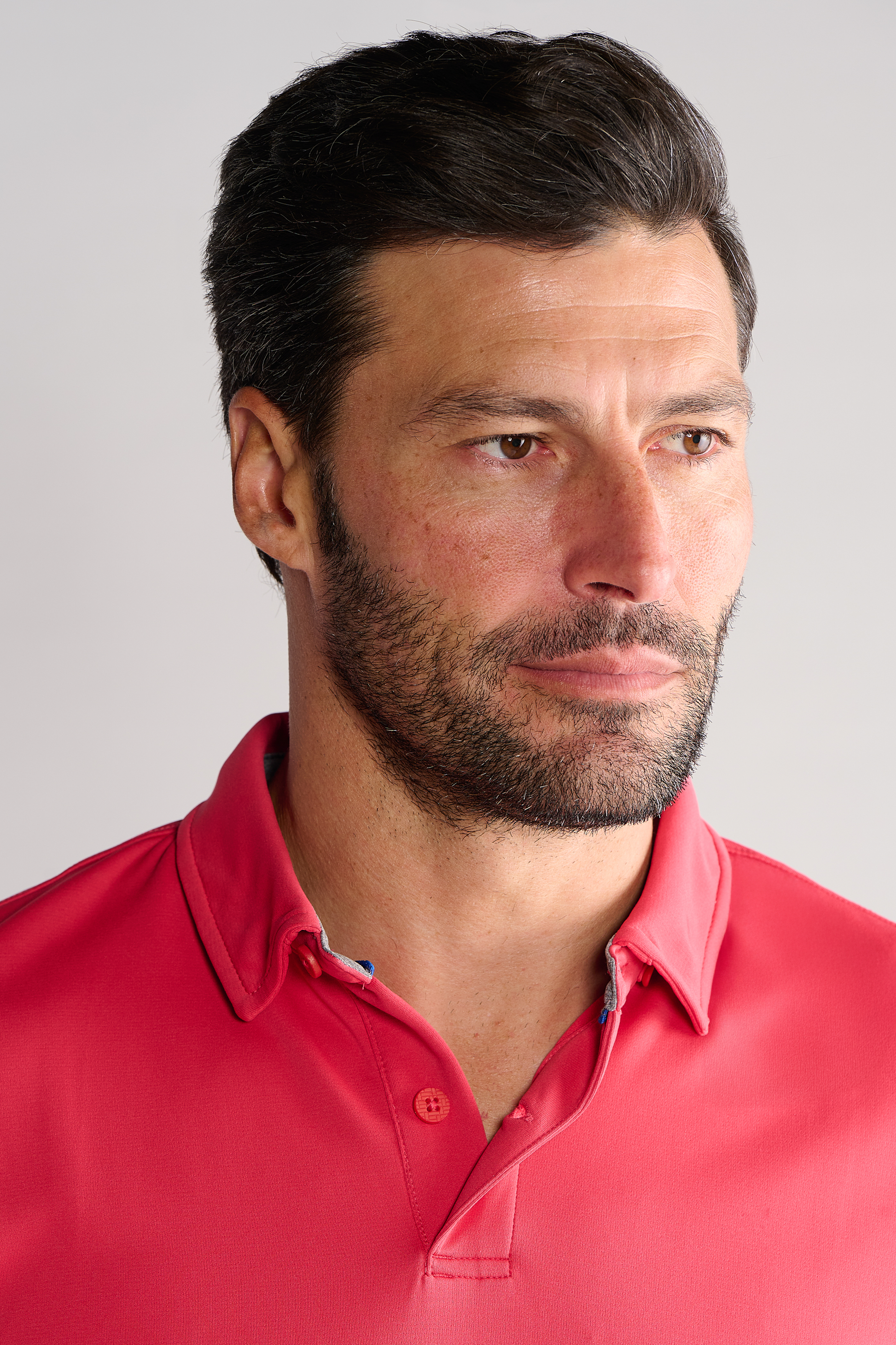 A man with short dark hair and a beard wears a Twillory Short Sleeve Polo Solid in red, looking slightly to the side with a neutral expression. A plain light background keeps focus on his face and upper torso.
