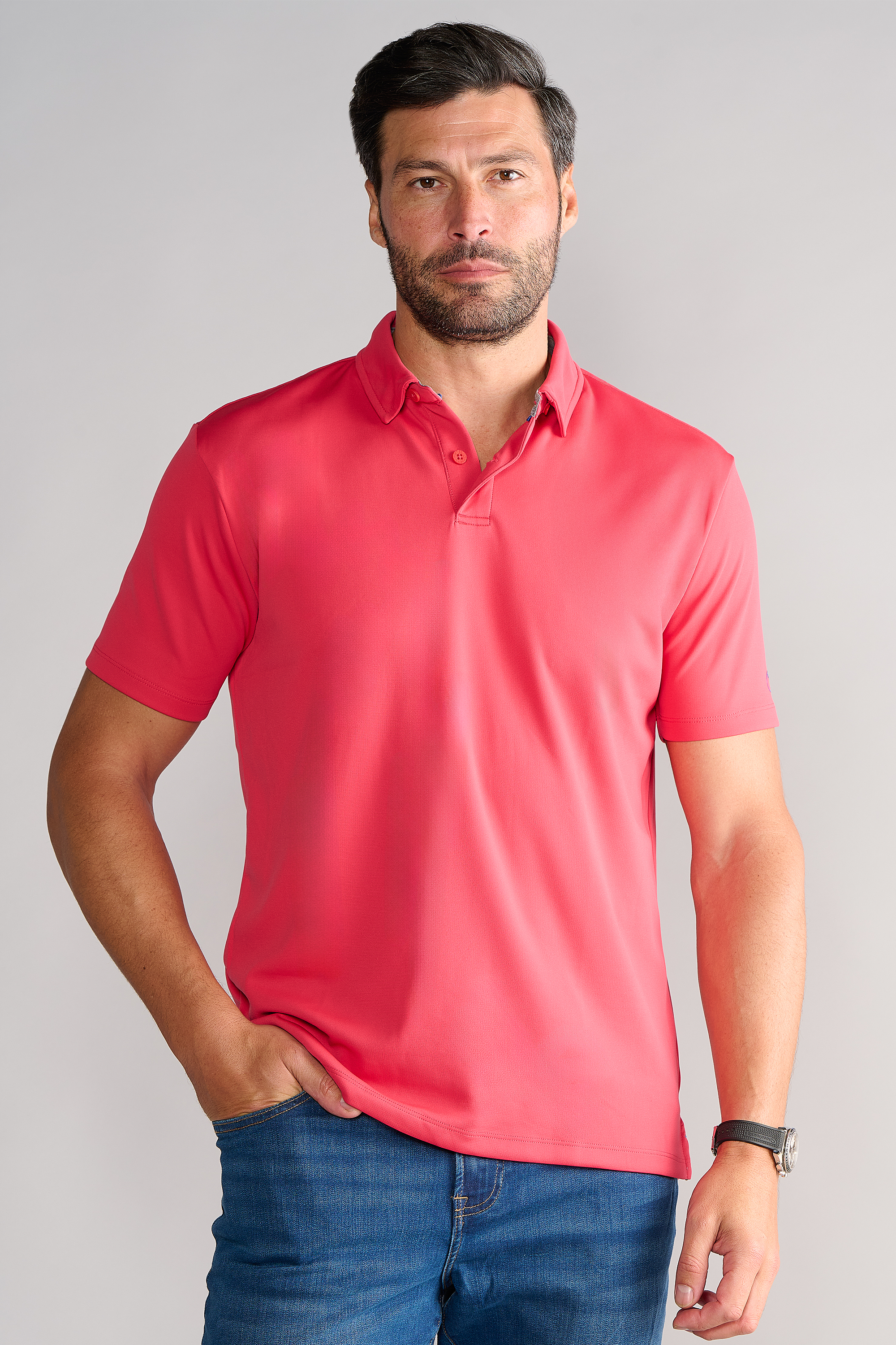 A man with dark hair and a beard stands against a light background, wearing the Twillory Short Sleeve Polo Solid in red and blue jeans, his left hand in his pocket and a watch on his wrist, looking directly at the camera.