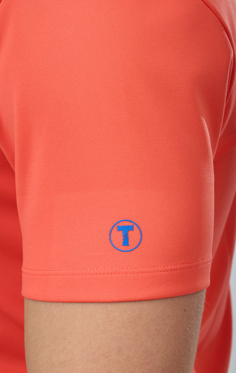 Close-up of a person’s upper arm in Twillory’s Short Sleeve Polo Solid // Salmon. A small blue circular logo with a stylized T appears near the sleeve hem, and the tailored fit with smooth fabric highlights comfort and style.