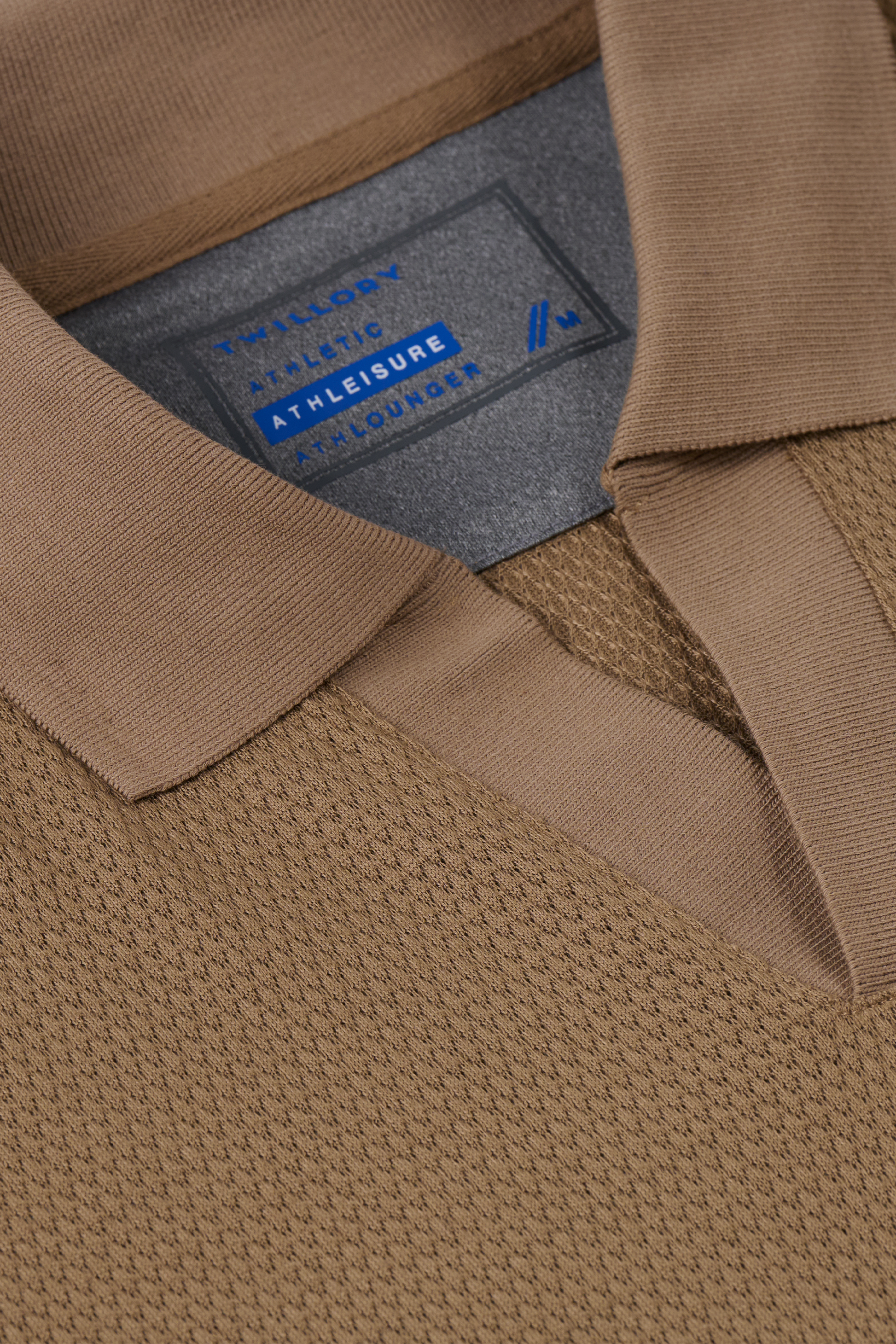 Close-up of the Twillory Short Sleeve Sweater Polo Solid in Sepia, featuring a textured waffle-like performance fabric, flat collar, and an ATHLEISURE label with blue and white text. The shirt is neatly folded.
