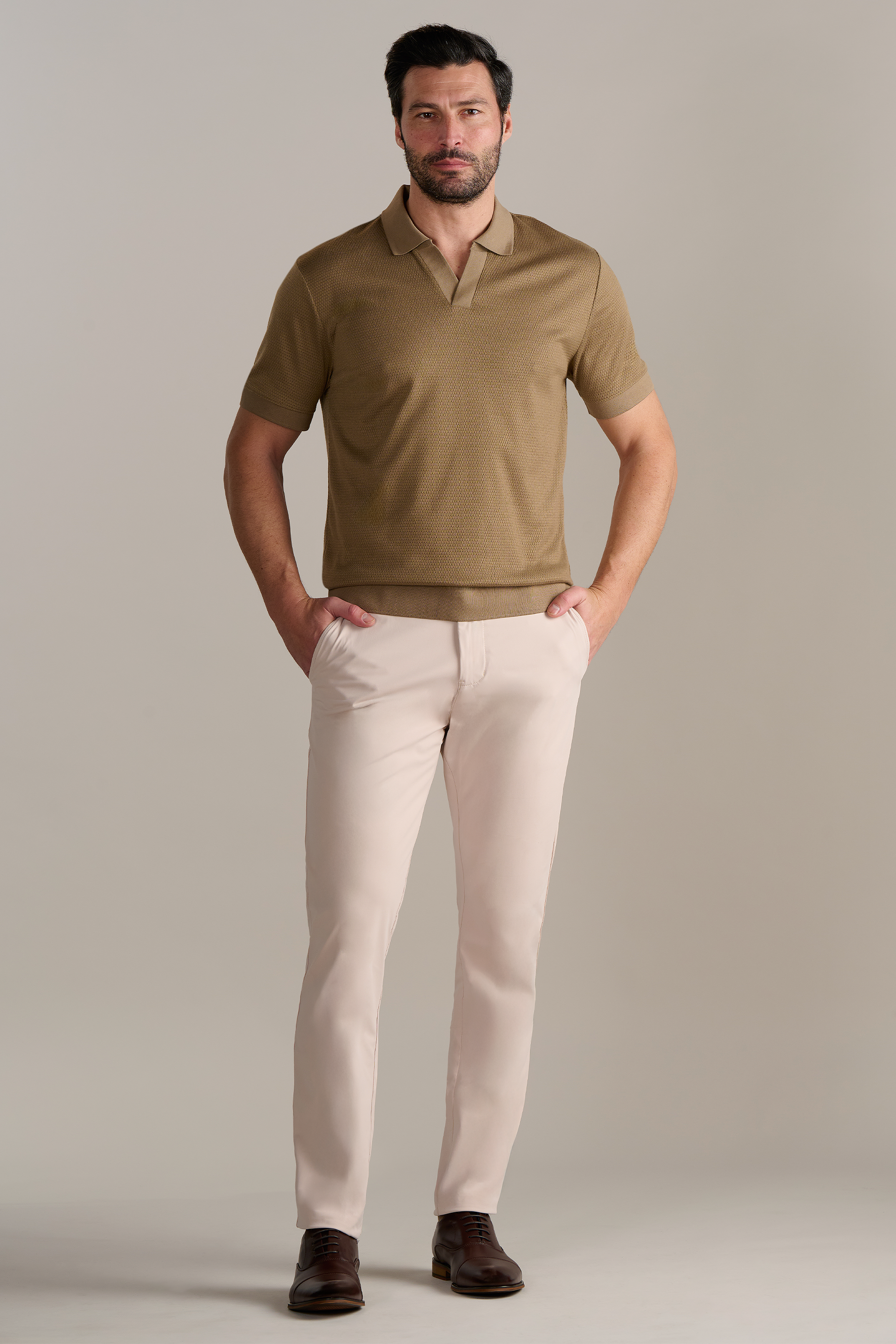 A man with dark hair and a beard stands against a light background, wearing Twillory’s Short Sleeve Sweater Polo Solid in Sepia tucked into slim-fit light beige pants and brown dress shoes, hands in pockets and looking relaxed.
