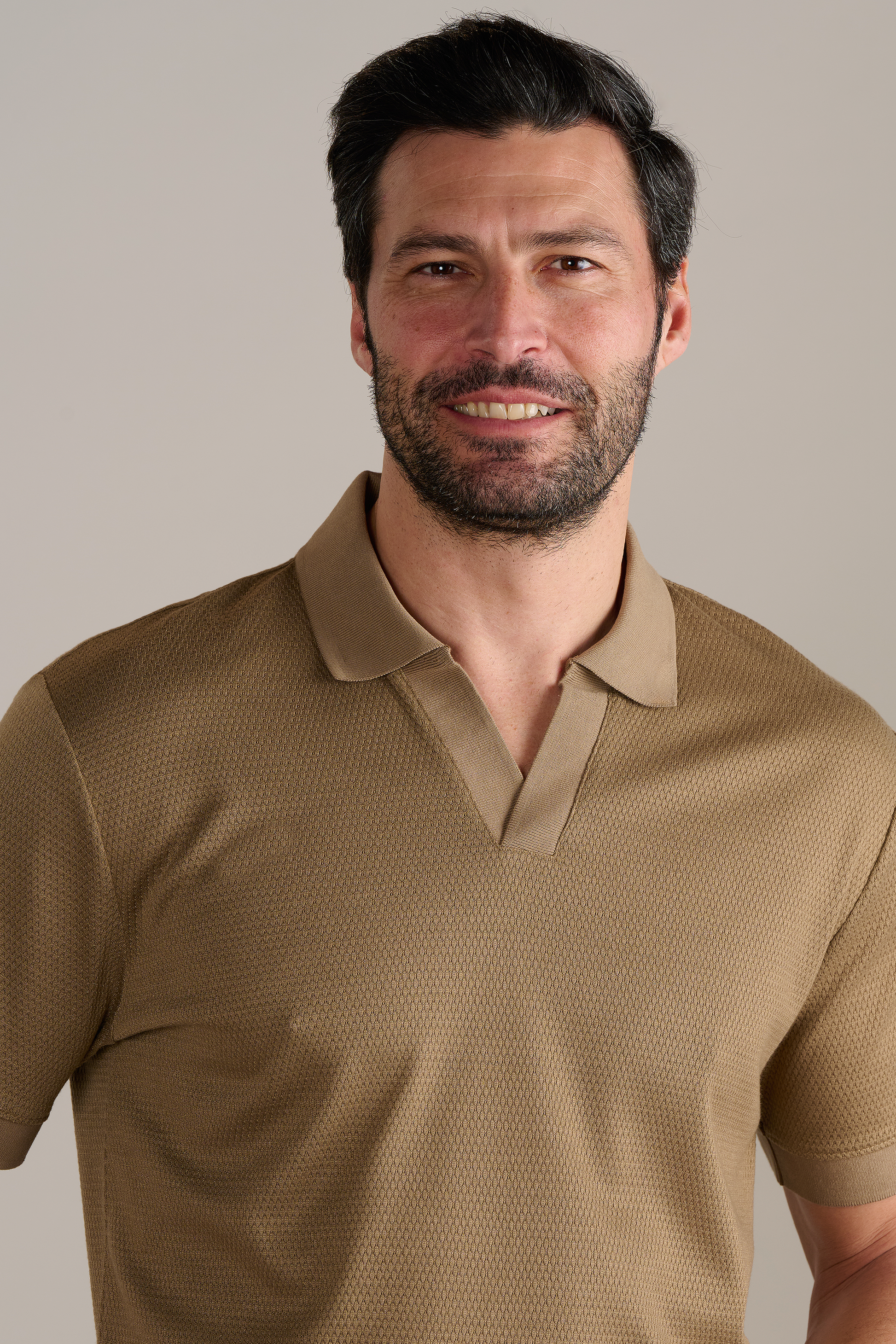 A man with short dark hair, a beard, and mustache gently smiles as he wears the Twillory Short Sleeve Sweater Polo Solid in Sepia. The tan textured knit shirt features a collar, with a neutral background highlighting his face and upper body.