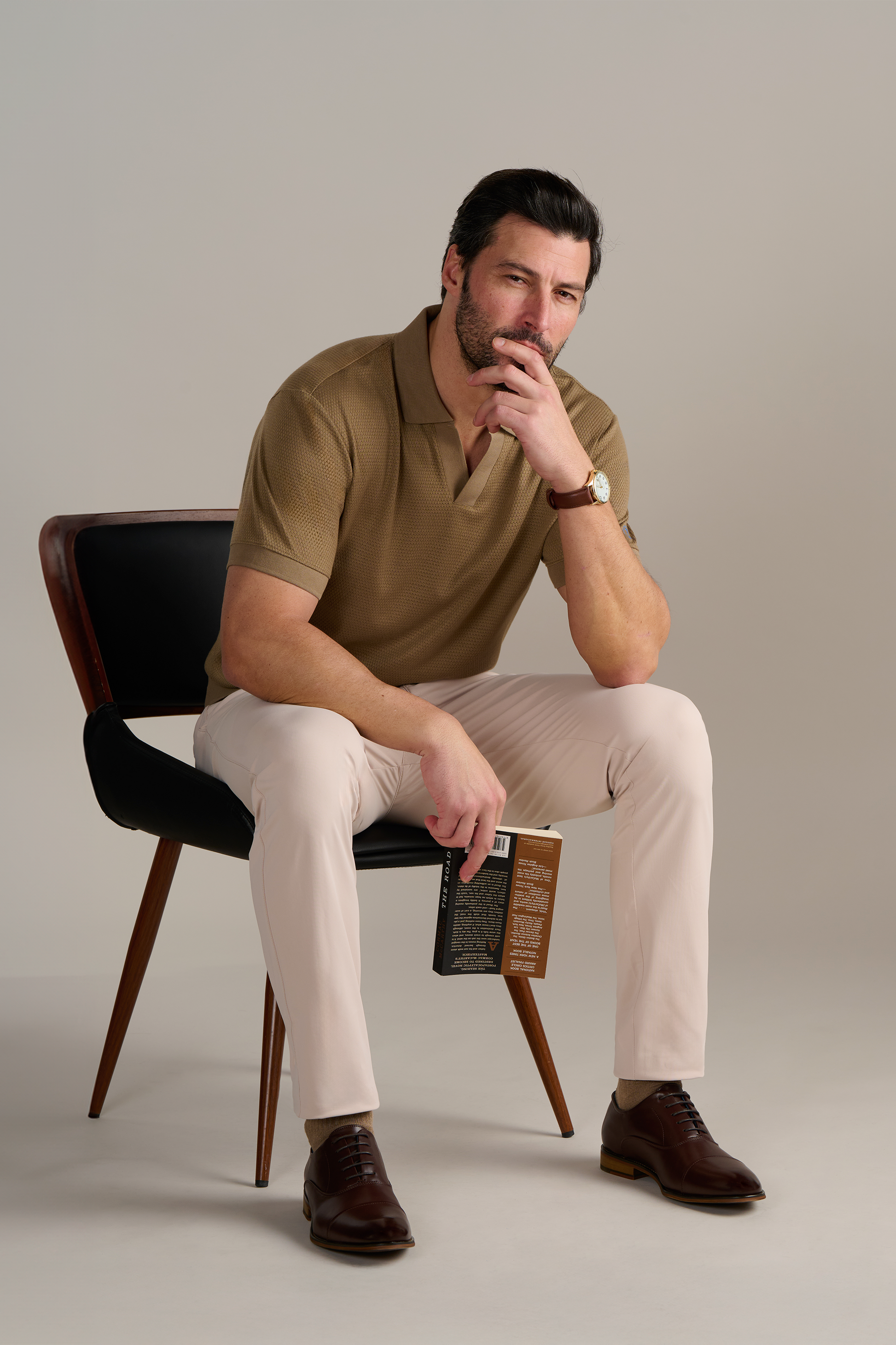 A man with short dark hair and a beard sits on a modern black chair wearing Twillory’s Short Sleeve Sweater Polo Solid in Sepia, light beige pants, brown leather shoes, and a wristwatch; he holds a closed book and rests his chin thoughtfully.