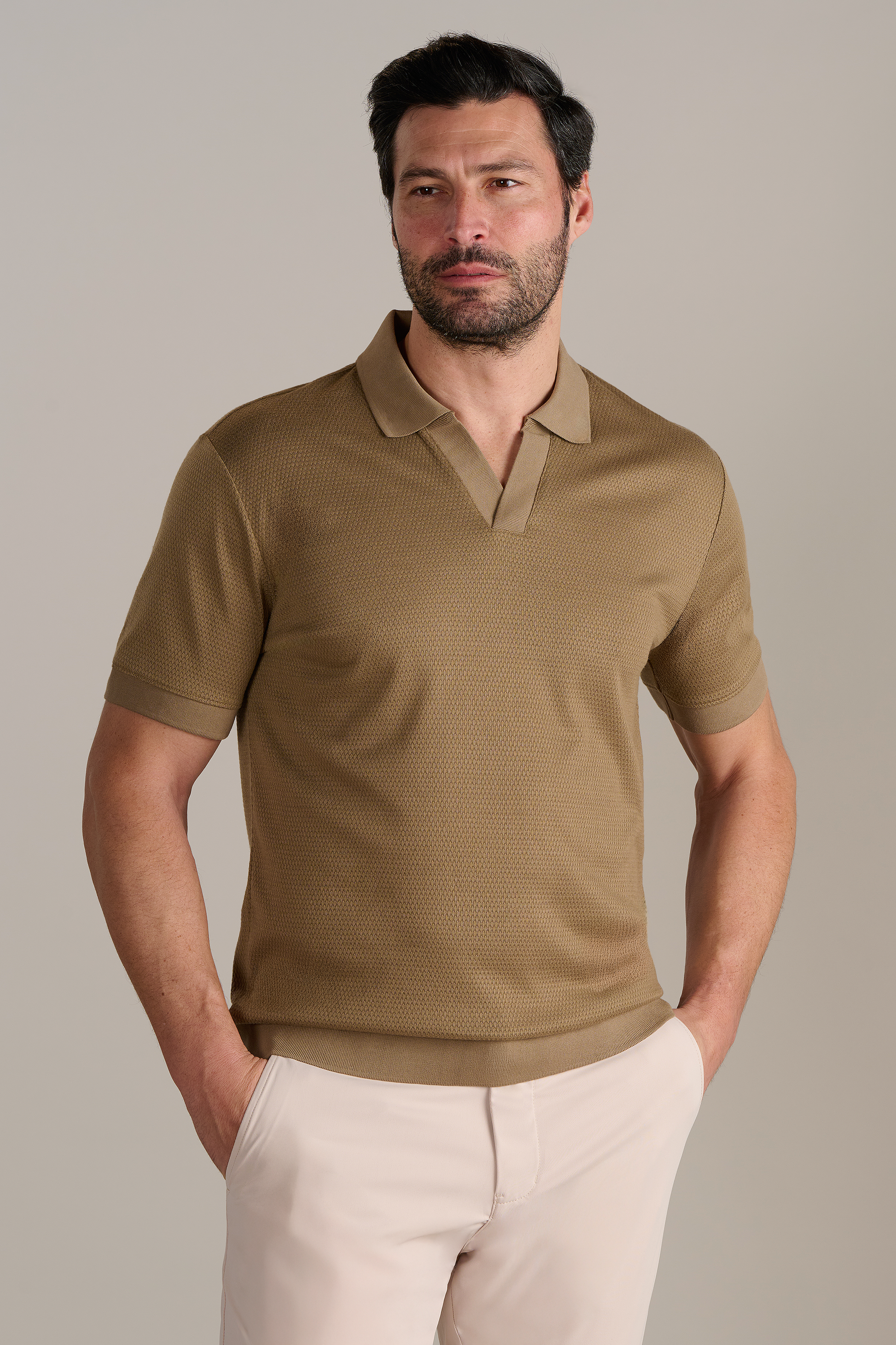 A man with short dark hair and a beard wears the Twillory Short Sleeve Sweater Polo Solid in Sepia and light beige pants, standing against a plain light gray background with his hands in his pockets, looking slightly to the side.