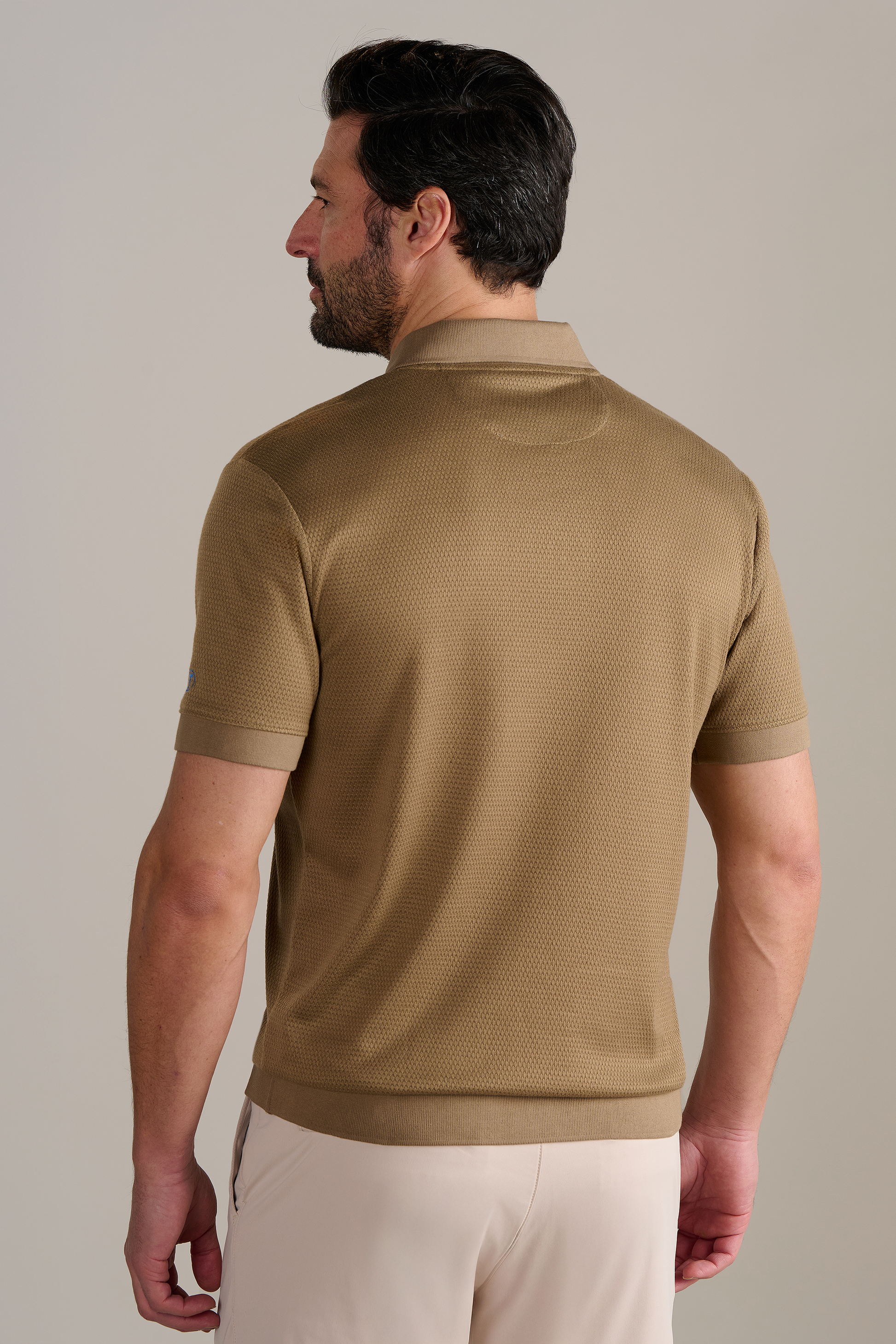 A man with light skin and dark, neatly styled hair faces away from the camera, wearing Twillory's Short Sleeve Sweater Polo Solid in Sepia and beige pants against a plain, neutral background.