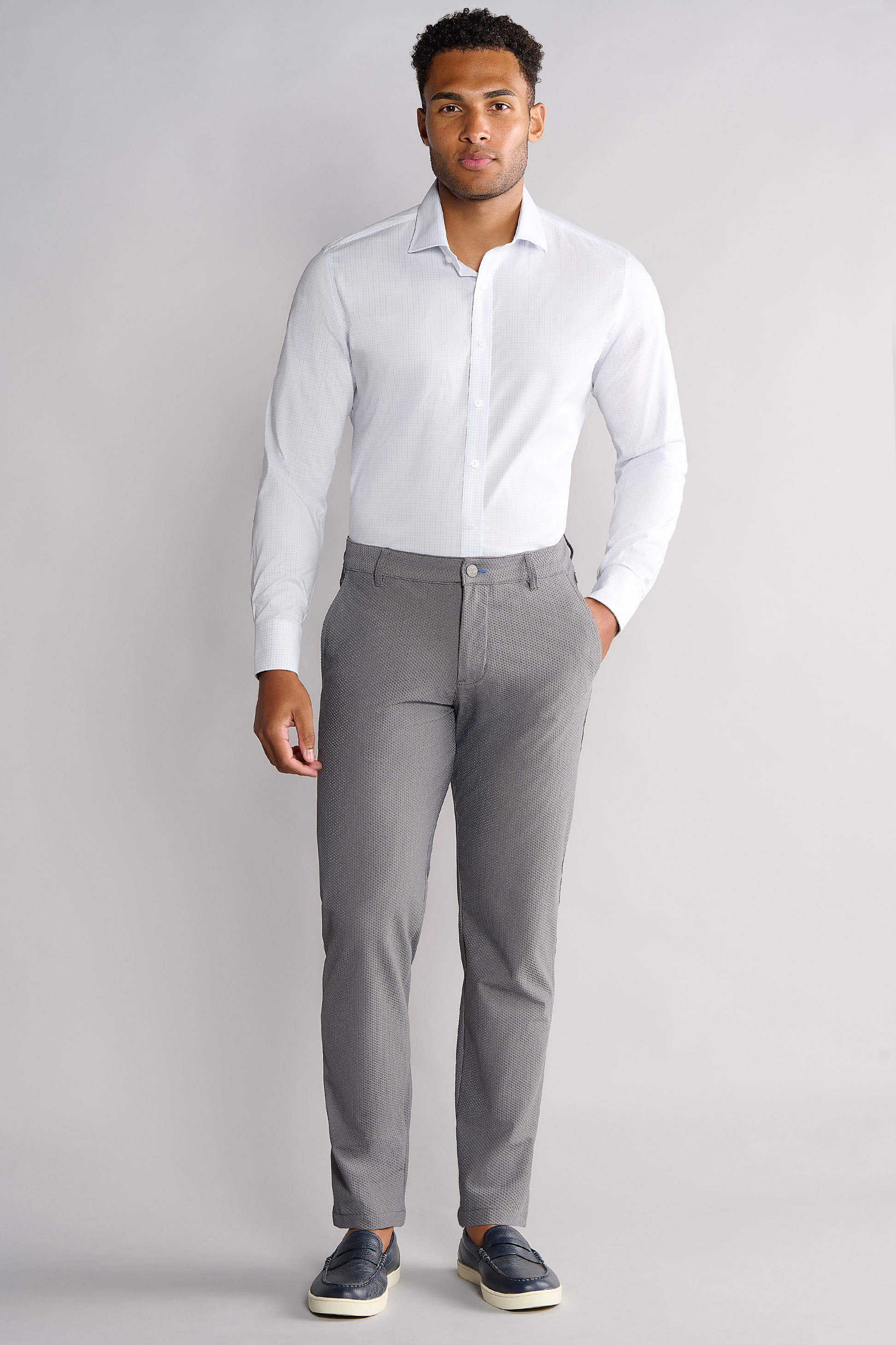 A man stands against a plain light grey background wearing the Twillory Leader Windowpane shirt in Sky Blue, paired with light grey slim-fit pants and dark slip-on shoes. One hand is in his pocket, and he looks straight ahead with a neutral expression.