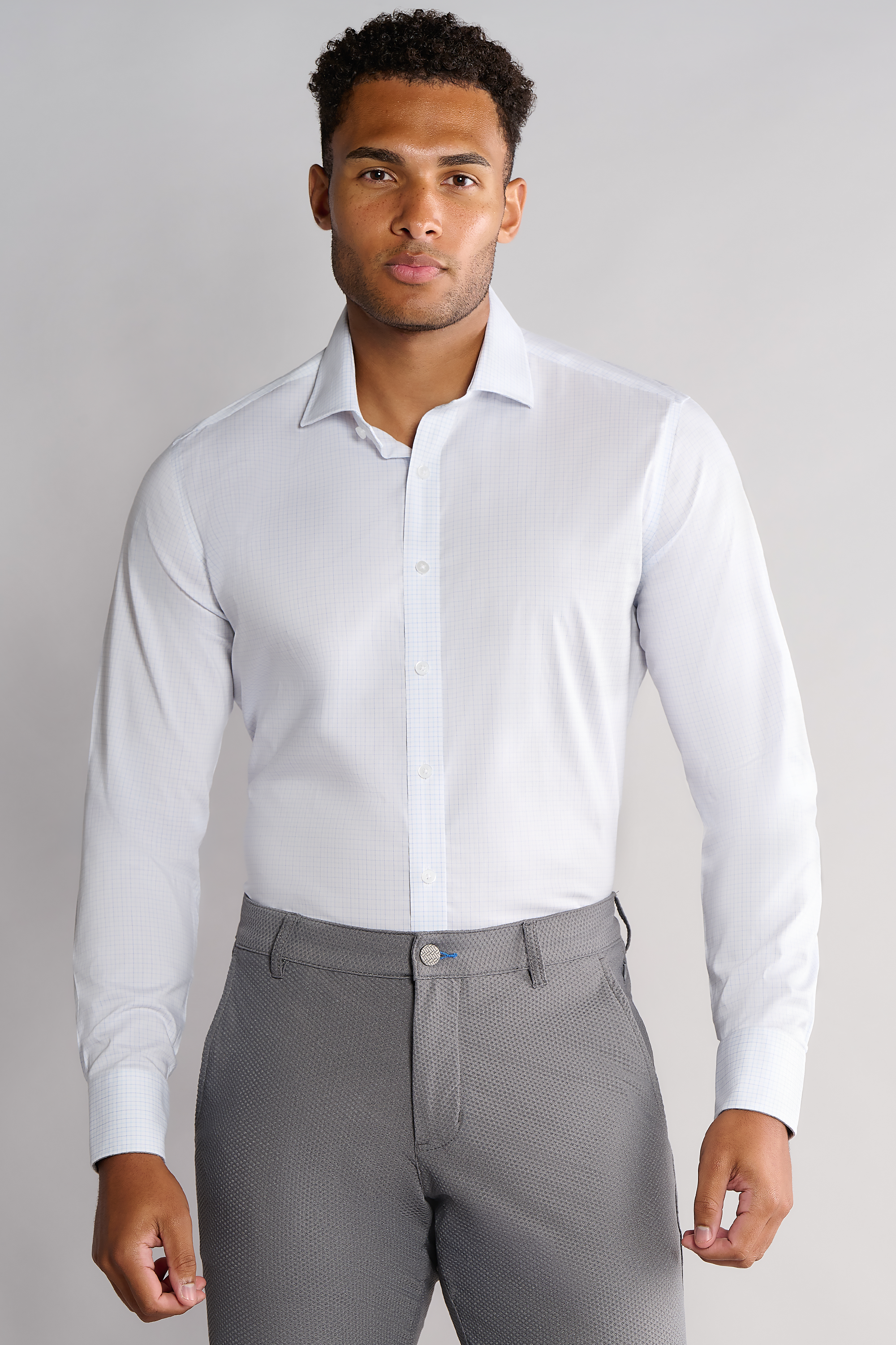 A man with short curly hair and a neutral expression stands facing forward against a plain background, wearing Twillory’s Leader Windowpane in Sky Blue—a non-iron performance shirt with a classic collar and subtle check—paired with gray dress pants.