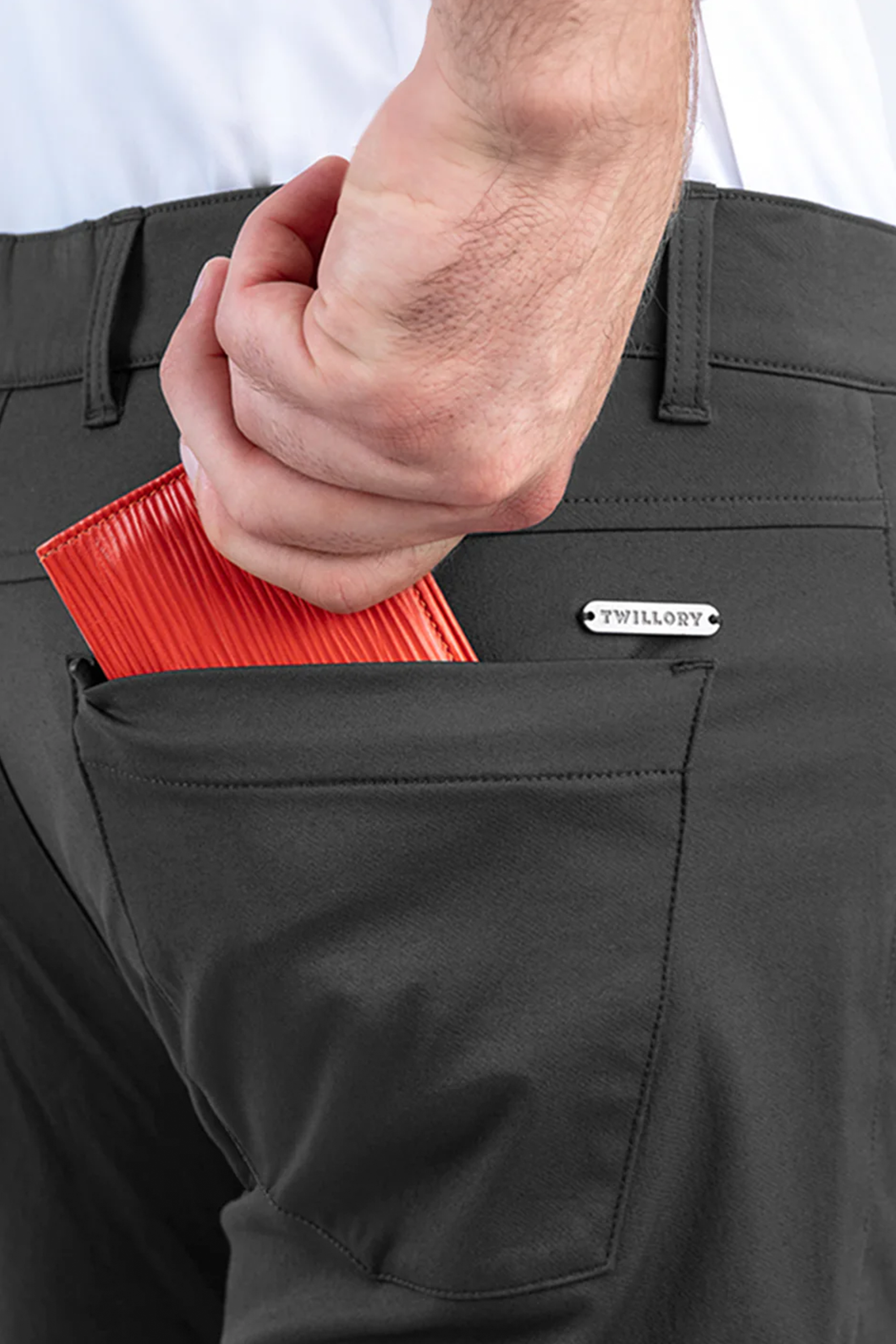 A close-up of a hand placing a red textured wallet into the back pocket of Twillory's Performance Five Pocket // Slate Grey pants, with the "Twillory" label above the pocket and a crisp white performance shirt neatly tucked in.