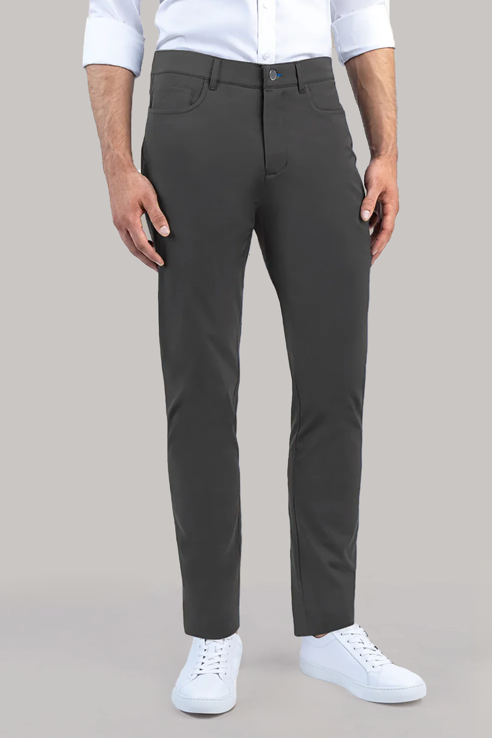 A person wears Twillory's Performance Five Pocket pants in Slate Grey, paired with a white long-sleeve shirt and white sneakers, standing against a plain light gray background. Only their lower torso and legs are shown.