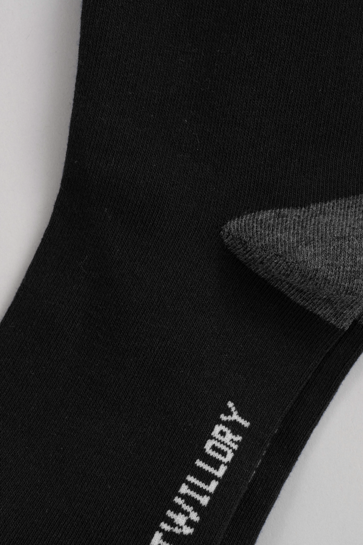 Close-up of Twillory’s “Black As Night” sock with a gray heel. The smooth, stretchy black fabric features part of a white word near the edge. Set against a plain light gray background, this could become your go-to sock.