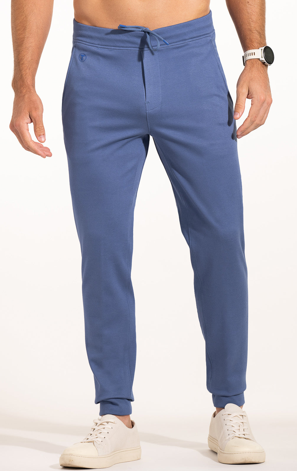 A man sports Twillory’s Athlounger Jogger—blue tapered sweatpants with drawstring waist, side-zip pocket, and side pockets—paired with white sneakers and a smartwatch, his shirtless torso visible against a light background.