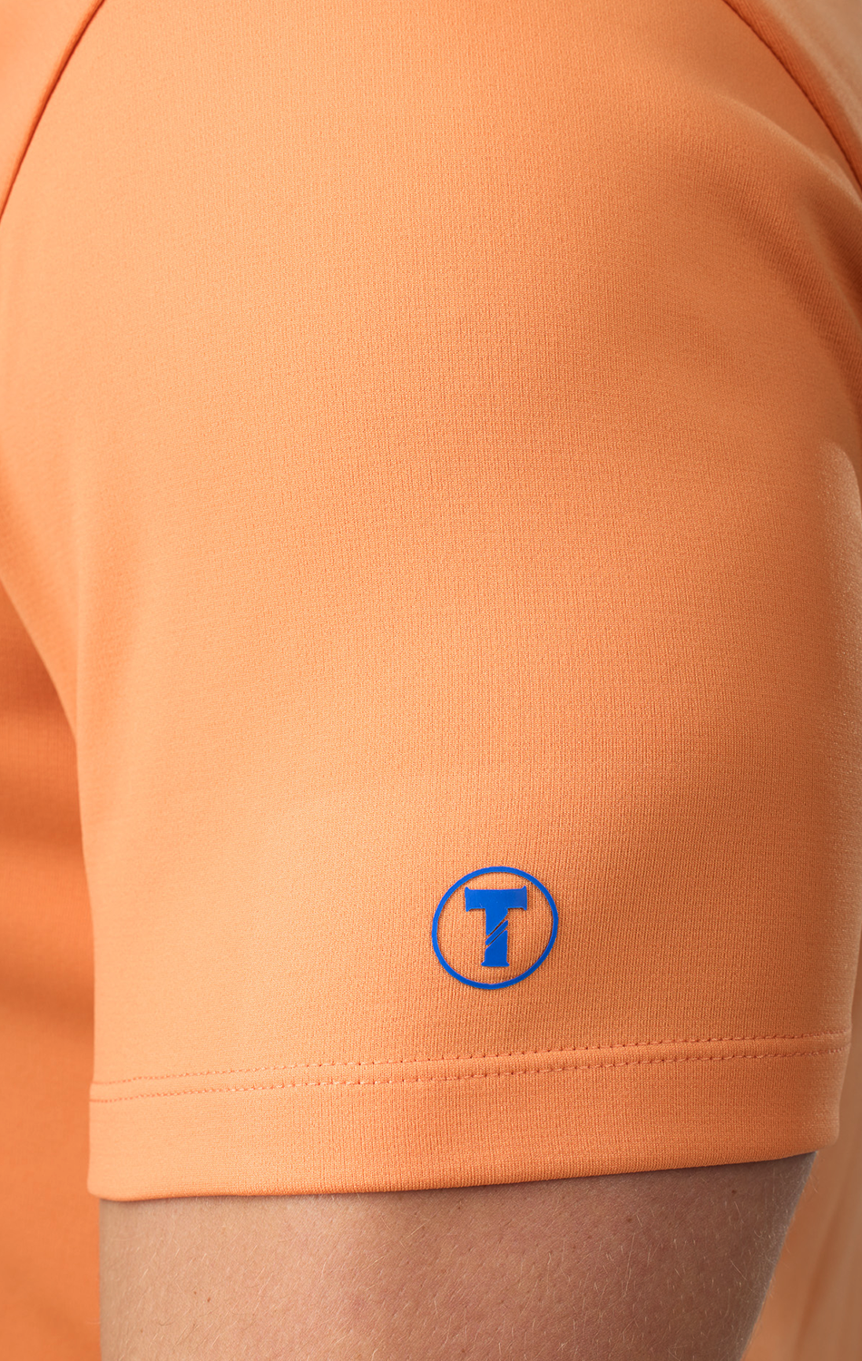 Close-up of a person wearing the Twillory Short Sleeve Polo Solid in Tangerine. This tailored-fit performance shirt features a blue circular “T” logo near the sleeve hem and smooth, moisture-wicking fabric fitted to the arm.