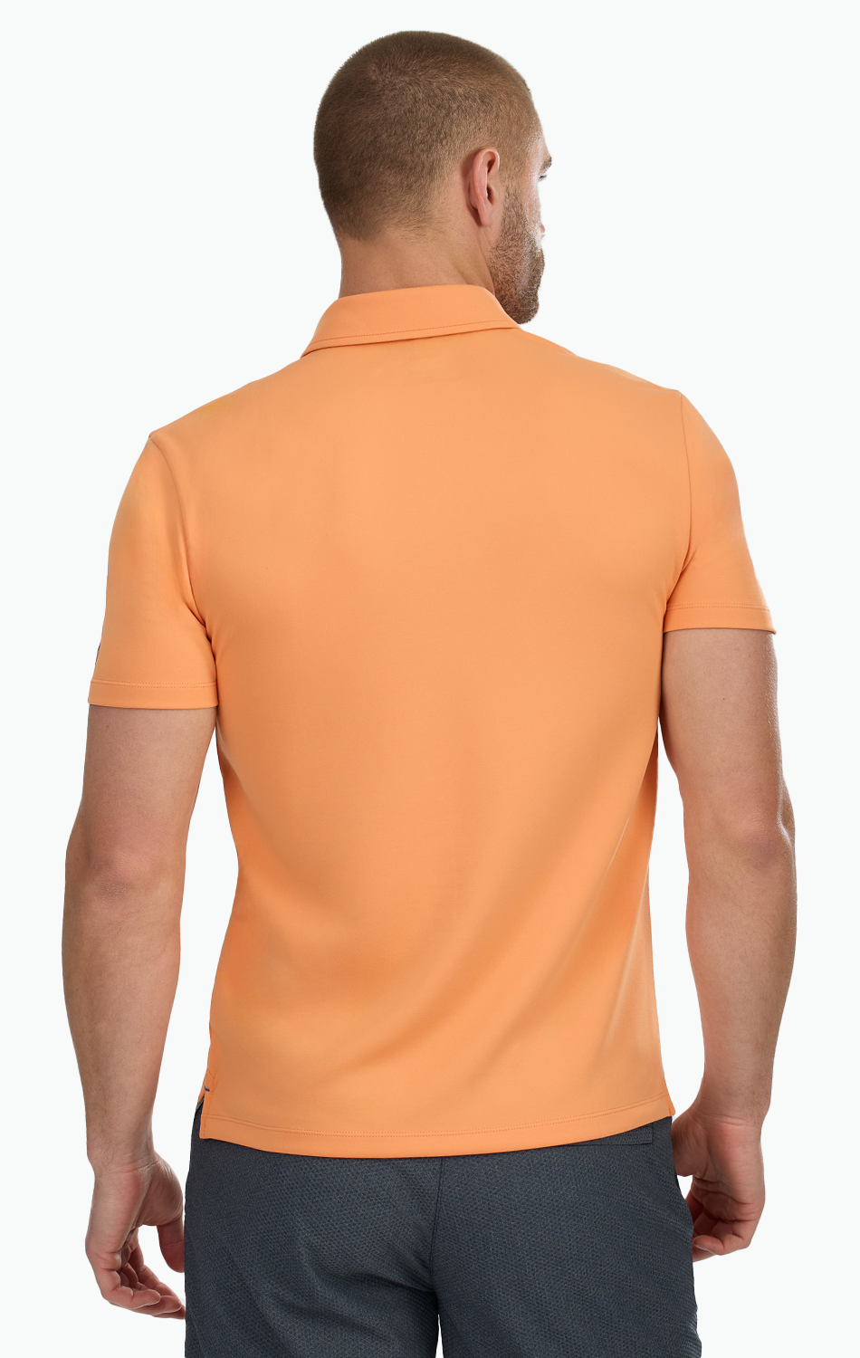 A man with short hair and a trimmed beard is seen from the back wearing Twillory's Short Sleeve Polo Solid in Tangerine with dark gray pants. His arms rest at his sides against a plain white background.