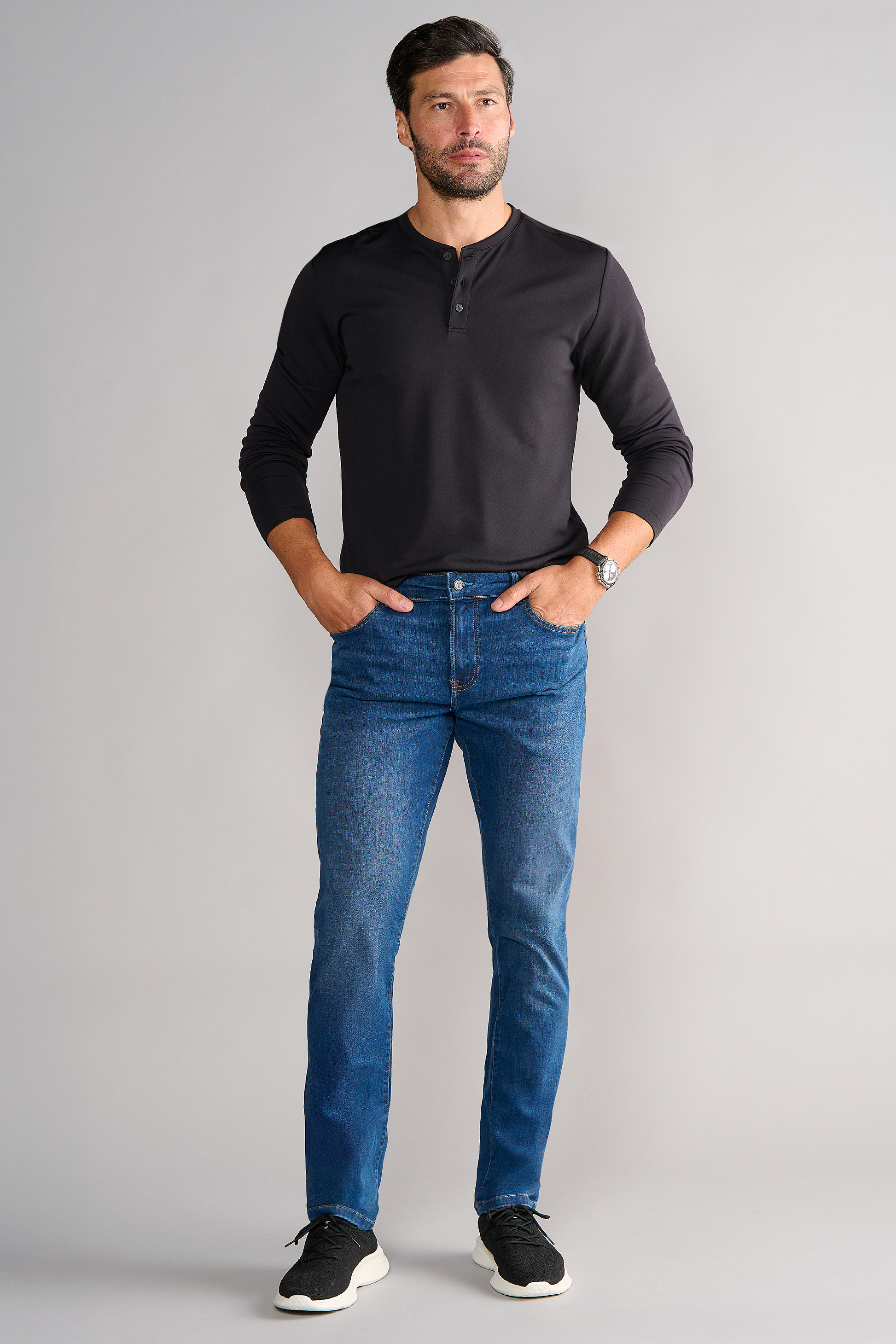 A man with short dark hair and a trimmed beard stands against a light gray background, wearing Twillory's Performance Denim in TRUE BLUE, a long-sleeve black henley shirt, and black sneakers. His hands are in his pockets and he has a neutral expression.