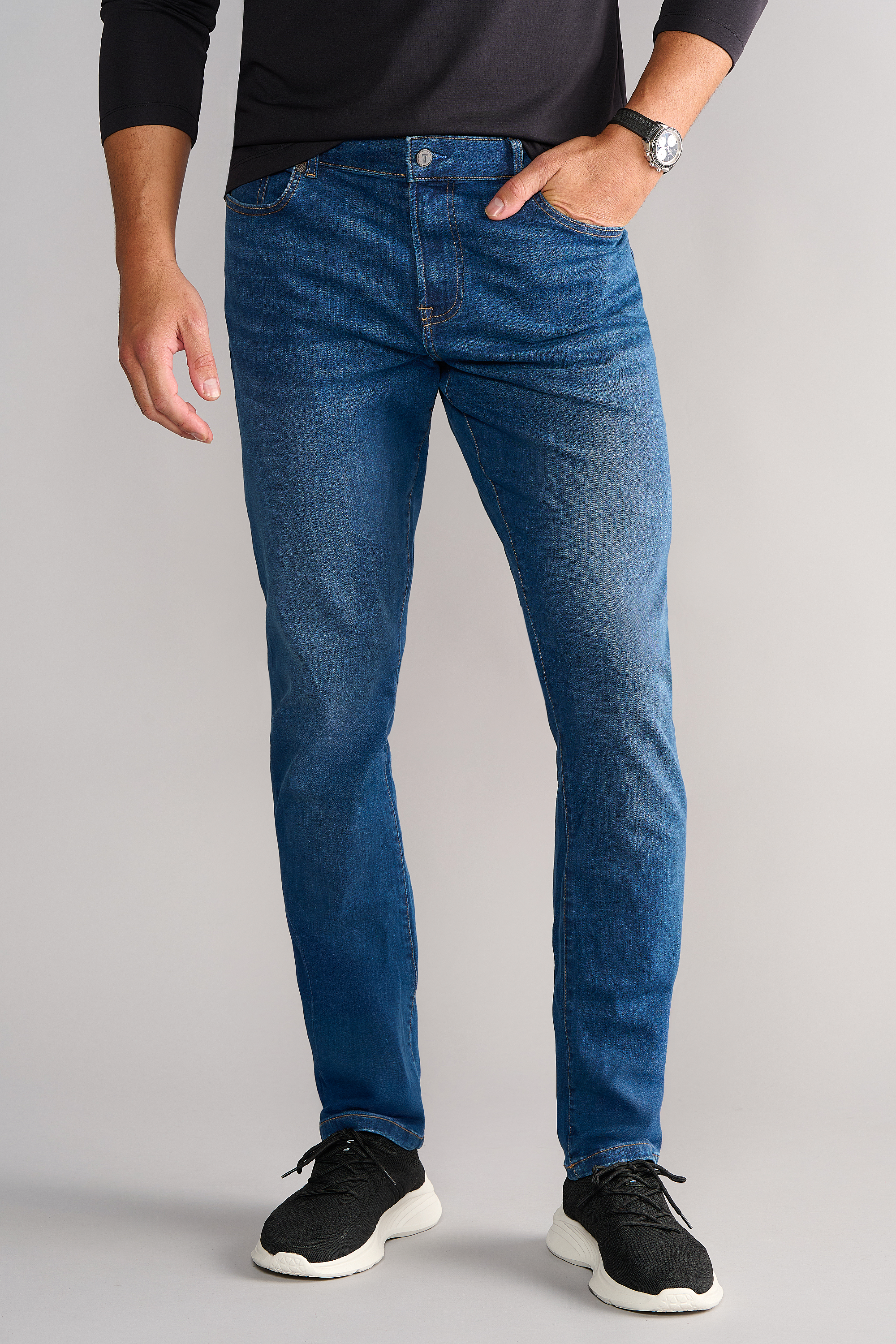 A person in a black long-sleeve shirt, Twillory Performance Denim // TRUE BLUE jeans, and black sneakers with white soles stands against a light gray background. They have their left hand in their pocket, showing a silver watch on their wrist.