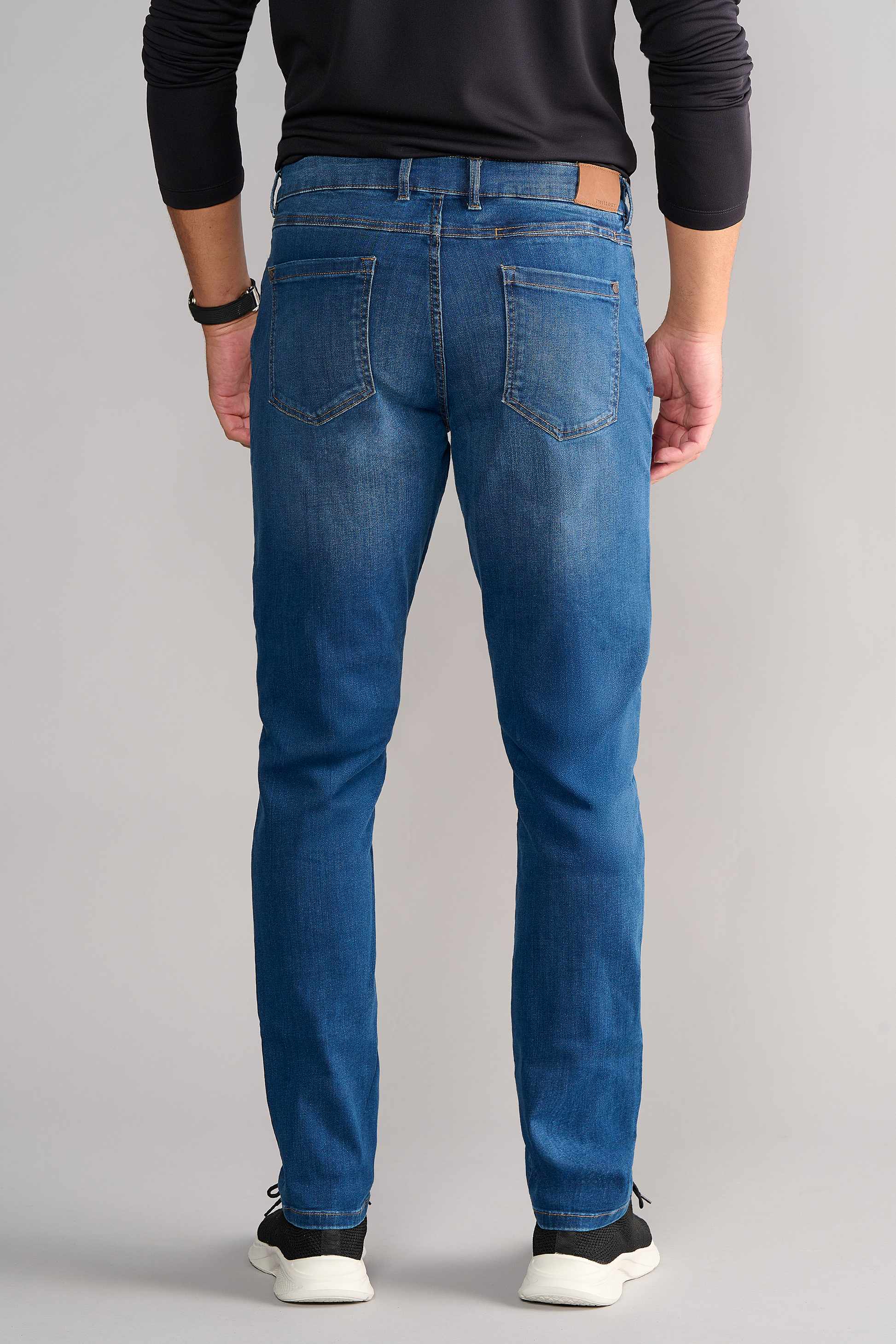 A person stands facing away in Twillory's Performance Denim // TRUE BLUE, featuring a tapered fit, two back pockets, and slight fading. Paired with a dark long-sleeve shirt, black watch, and black-and-white sneakers against a light gray background.