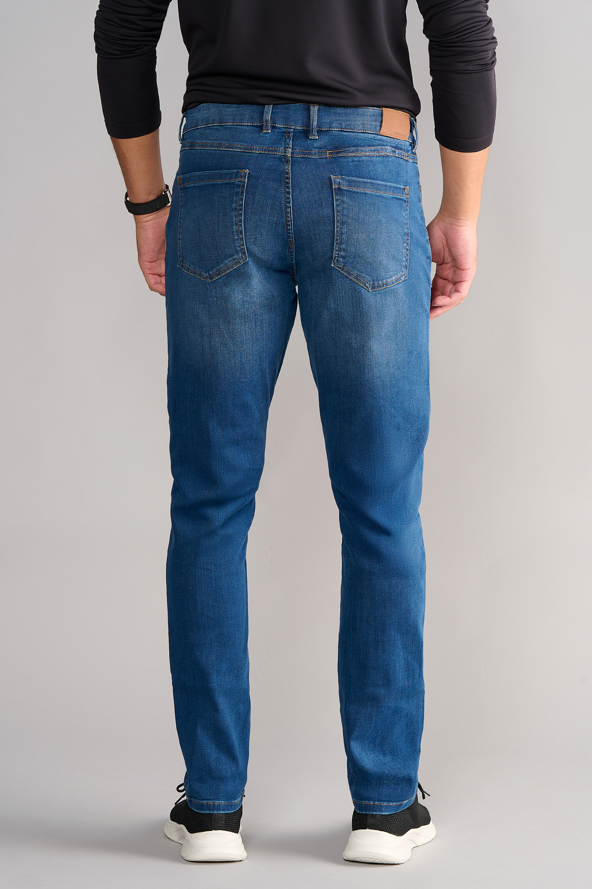 A person stands facing away in Twillory's Performance Denim // TRUE BLUE, featuring a tapered fit, two back pockets, and slight fading. Paired with a dark long-sleeve shirt, black watch, and black-and-white sneakers against a light gray background.