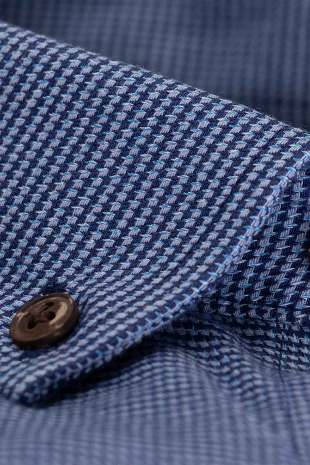 Close-up of Twillory's untuck(able) Blue Puppytooth shirt, featuring a blue and white puppytooth pattern, folded collar, dark brown button, and non-iron stretch fabric that showcases its detailed weave and texture.