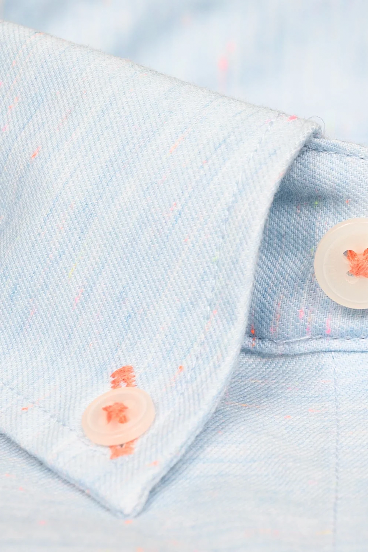 Close-up of the untuck(able) Caribbean Melange shirt cuff by Twillory: light blue non-iron stretch fabric with faint multicolored specks, a diagonal weave, and two white buttons sewn with orange thread highlight its fine details.