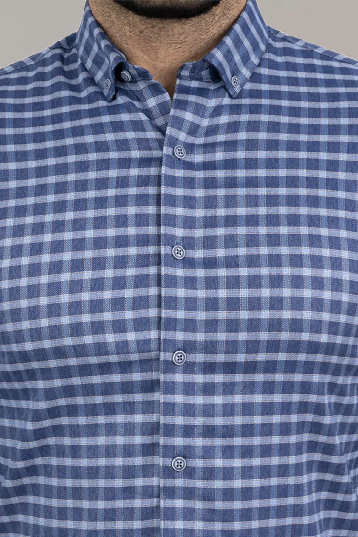 A close-up shows a person wearing Twillory’s untuck(able) Denim Windowpane shirt, featuring a blue and white checkered grid pattern with a button-down collar; only the chest and neck are visible, face not shown.