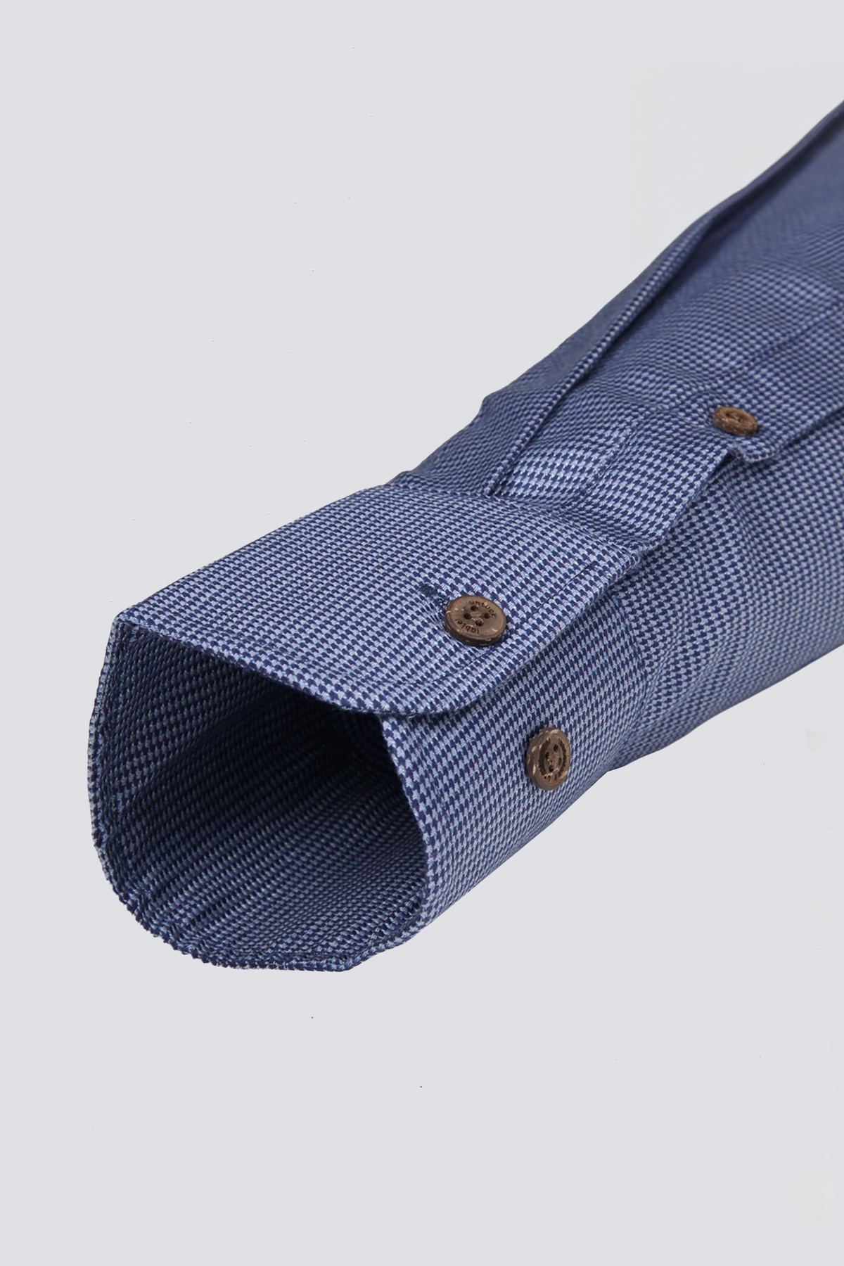 Close-up of the Twillory untuck(able) Blue Puppytooth shirt cuff, featuring a subtle puppytooth pattern, non-iron stretch fabric, two brown buttons, and visible stitching, shown against a plain light gray background.