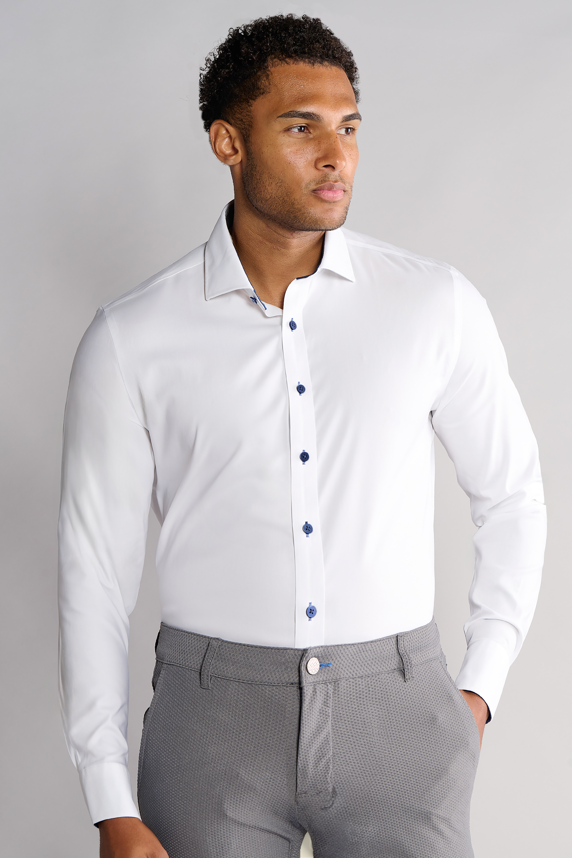 A man stands against a light gray background wearing Twillory's Leader Contrast // Navy, a fitted moisture-wicking performance shirt with blue buttons, and light gray dress pants. He looks right with a neutral expression, left hand at his side and right hand in his pocket.