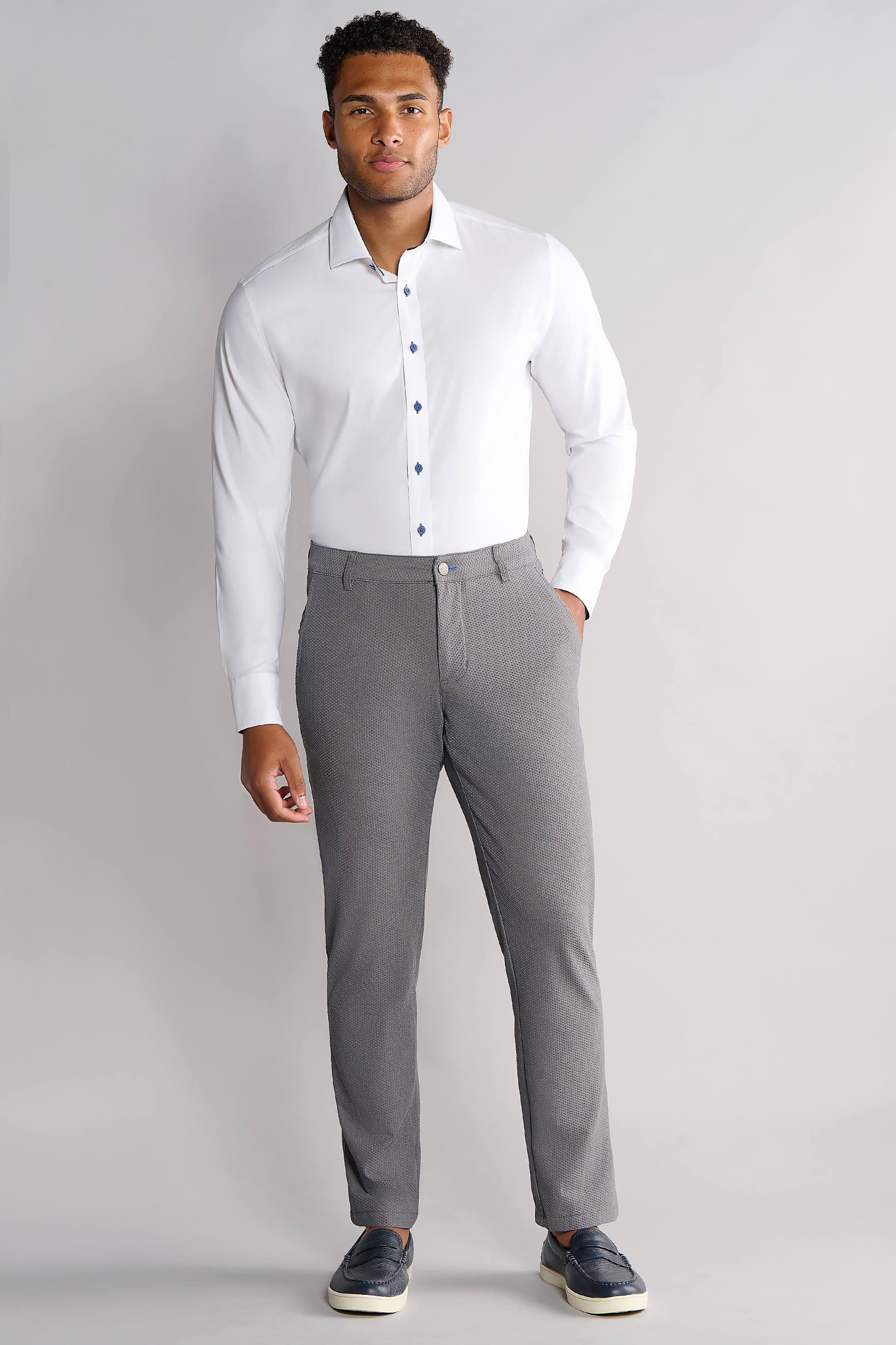 A man stands against a light gray background wearing Twillory's Leader Contrast // Navy, a white 4-way stretch performance shirt with dark buttons, paired with slim-fit light gray trousers and dark blue slip-on shoes.