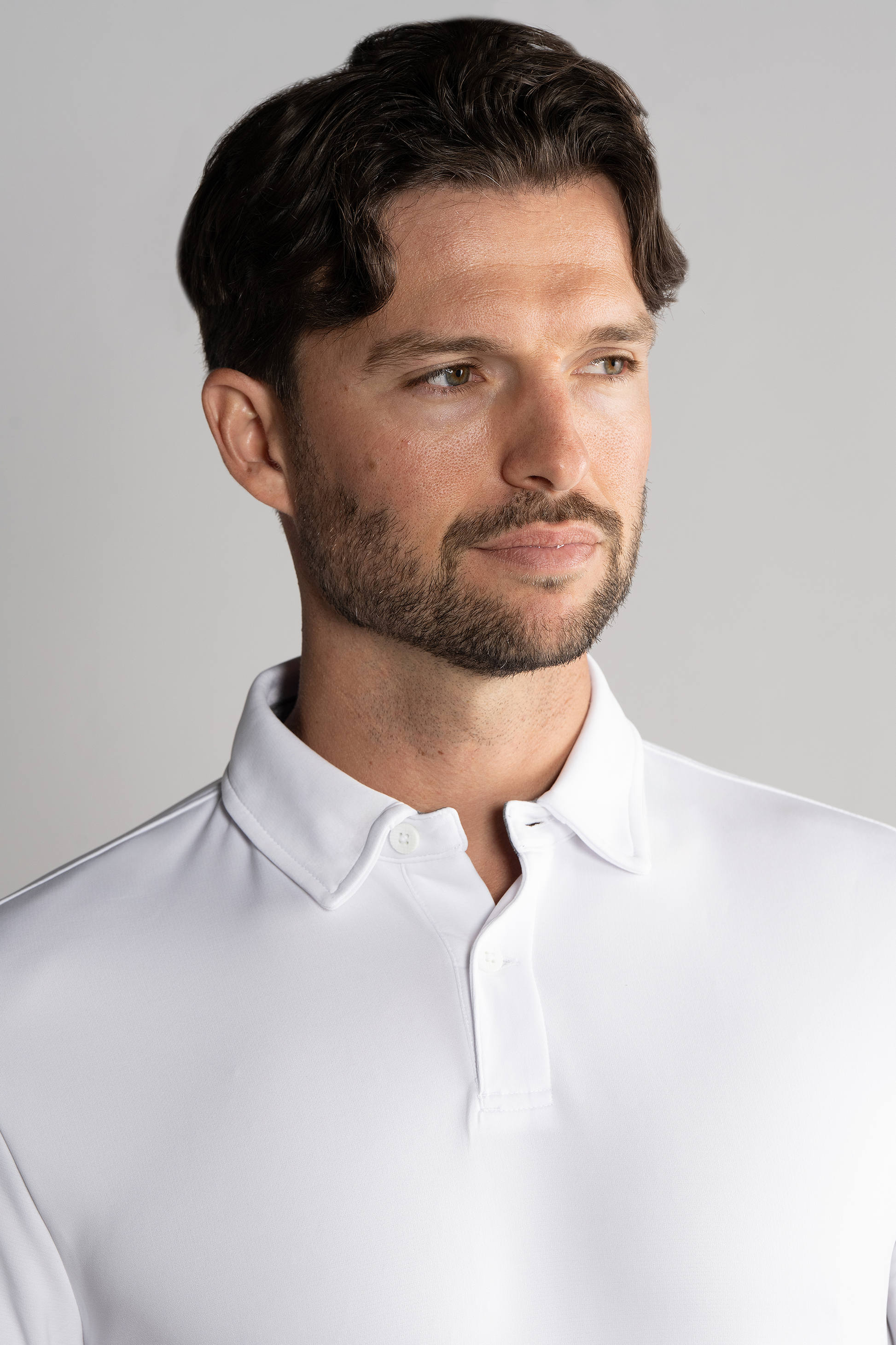 A man with short, wavy brown hair and a trimmed beard wears the Twillory Short Sleeve Polo Solid in White. He looks slightly right with a neutral expression against a clean, light gray background.