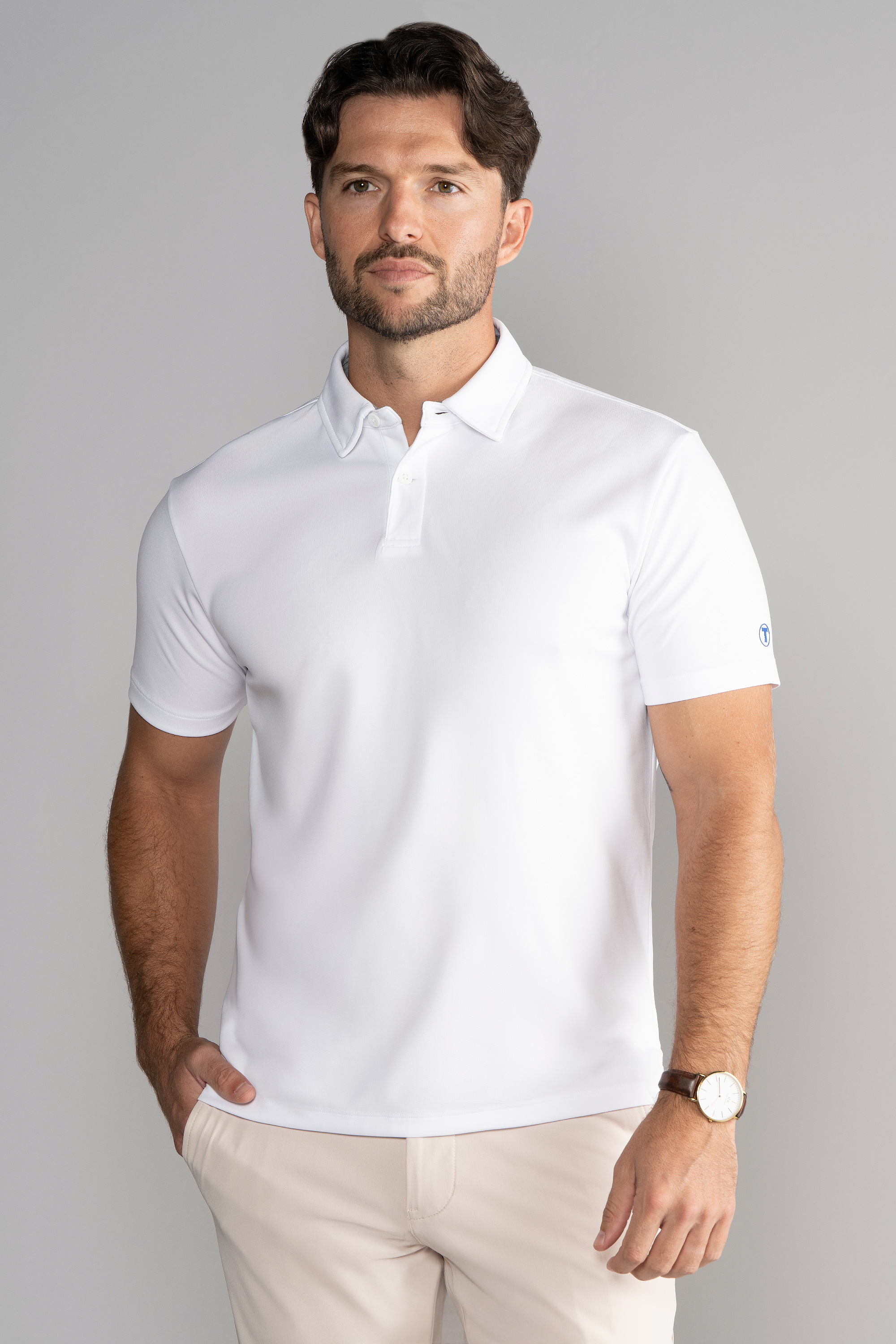 A man with wavy brown hair and a short beard stands against a plain gray background, wearing the Twillory Short Sleeve Polo Solid in White, light beige pants, and a brown watch, with one hand in his pocket and a neutral expression.