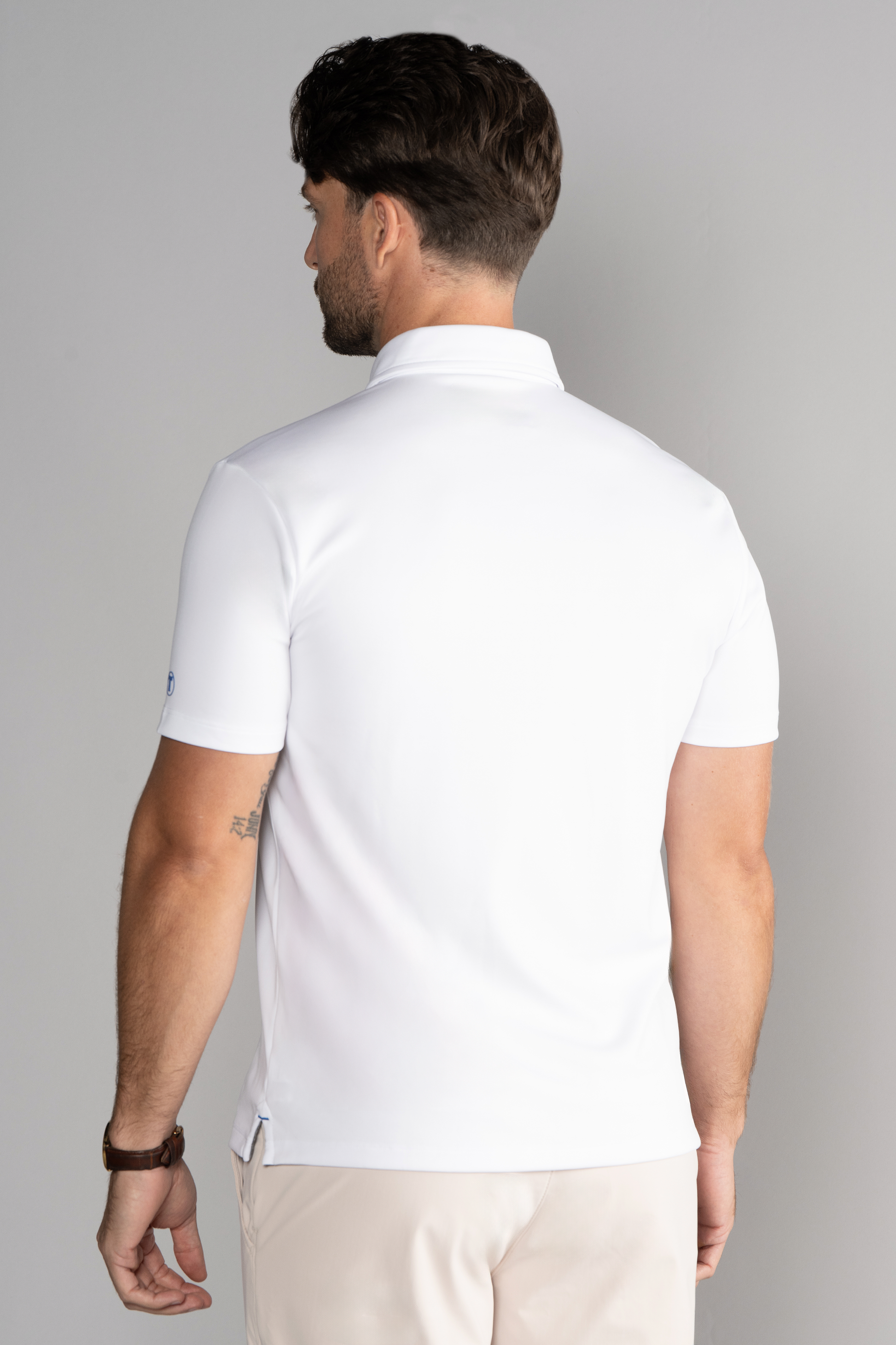 A man with neatly styled dark hair wears the Twillory Short Sleeve Polo Solid in White and light beige pants, accessorized with a watch on his left wrist. He faces away from the camera against a plain light gray background.